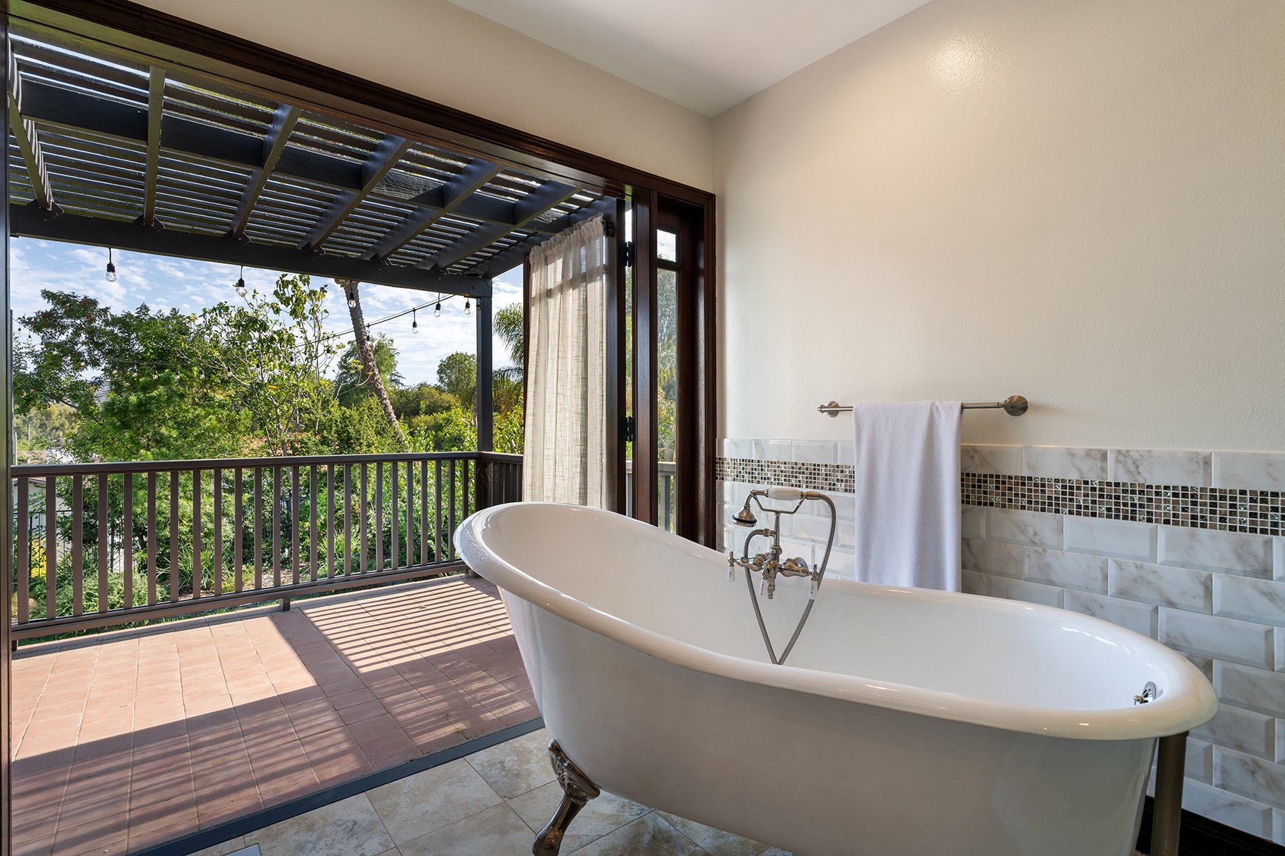 A bathroom with a clawfoot bathtub facing a balcony with a view of trees and blue sky, featuring sliding glass doors with curtains, a decorative tile wall, a towel rack with a white towel, and outdoor string lights.