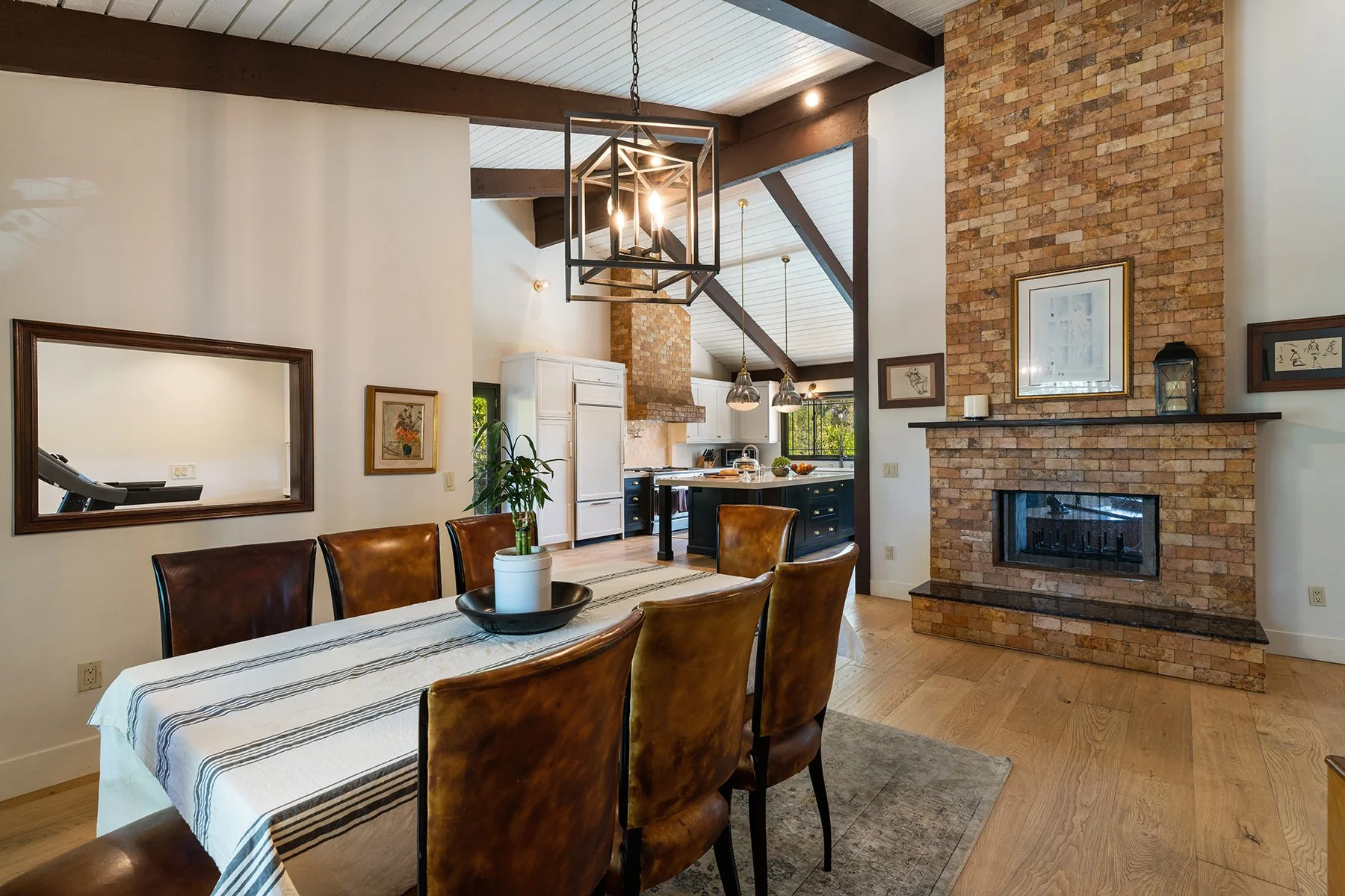 A dining room with a view of the kitchen featuring a brick fireplace, wooden beams, and modern lighting fixtures. The dining table has a striped tablecloth and leather chairs.