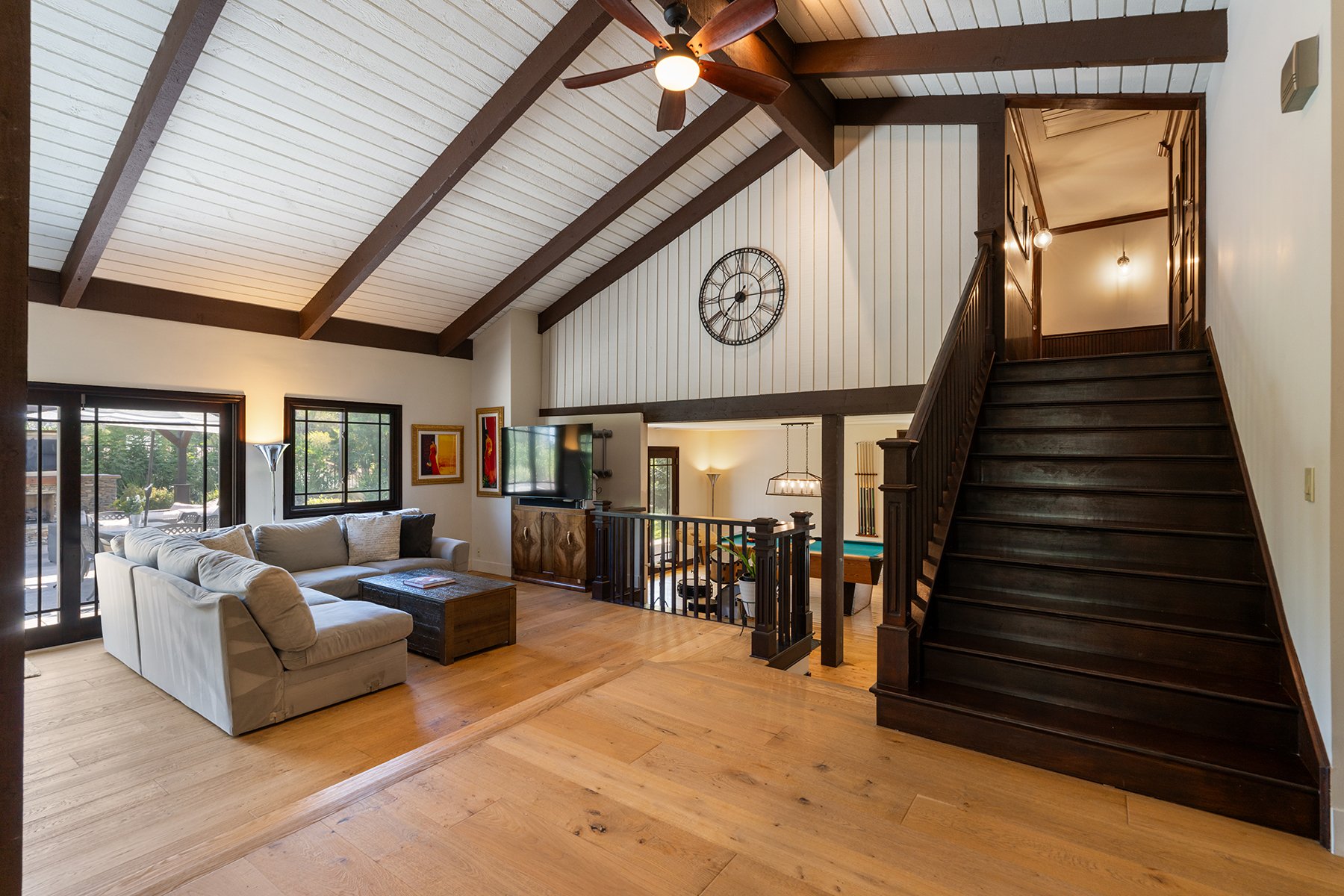 Living room with high vaulted ceiling, exposed dark wood beams, ceiling fan, large windows, beige sectional sofa, wooden coffee table, wall-mounted television, artwork, and a staircase leading to an upper level.