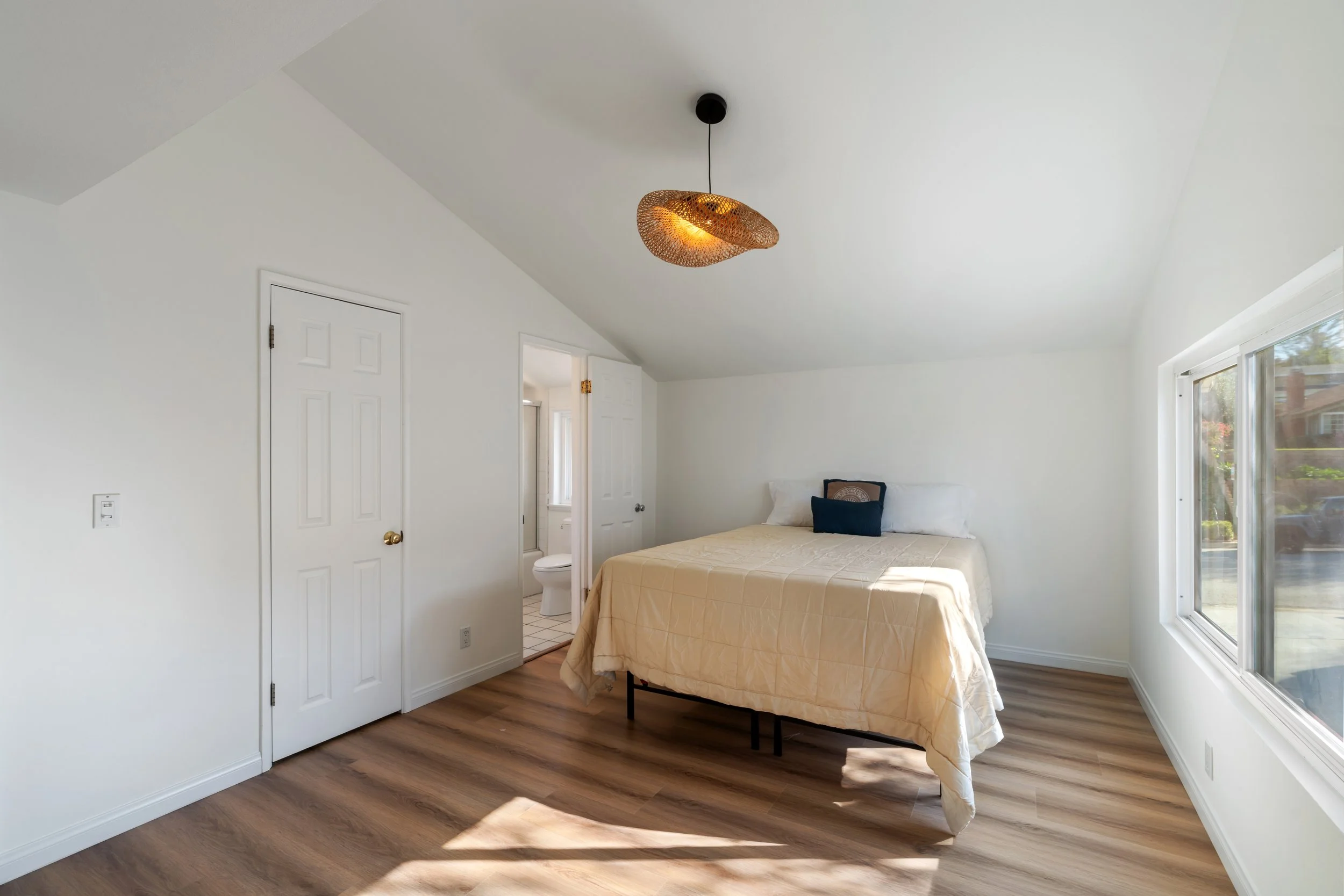 Bedroom with a bed featuring a beige quilt, white pillows, and a decorative pillow. Large window on the right, white walls, wood flooring, and a ceiling light fixture.