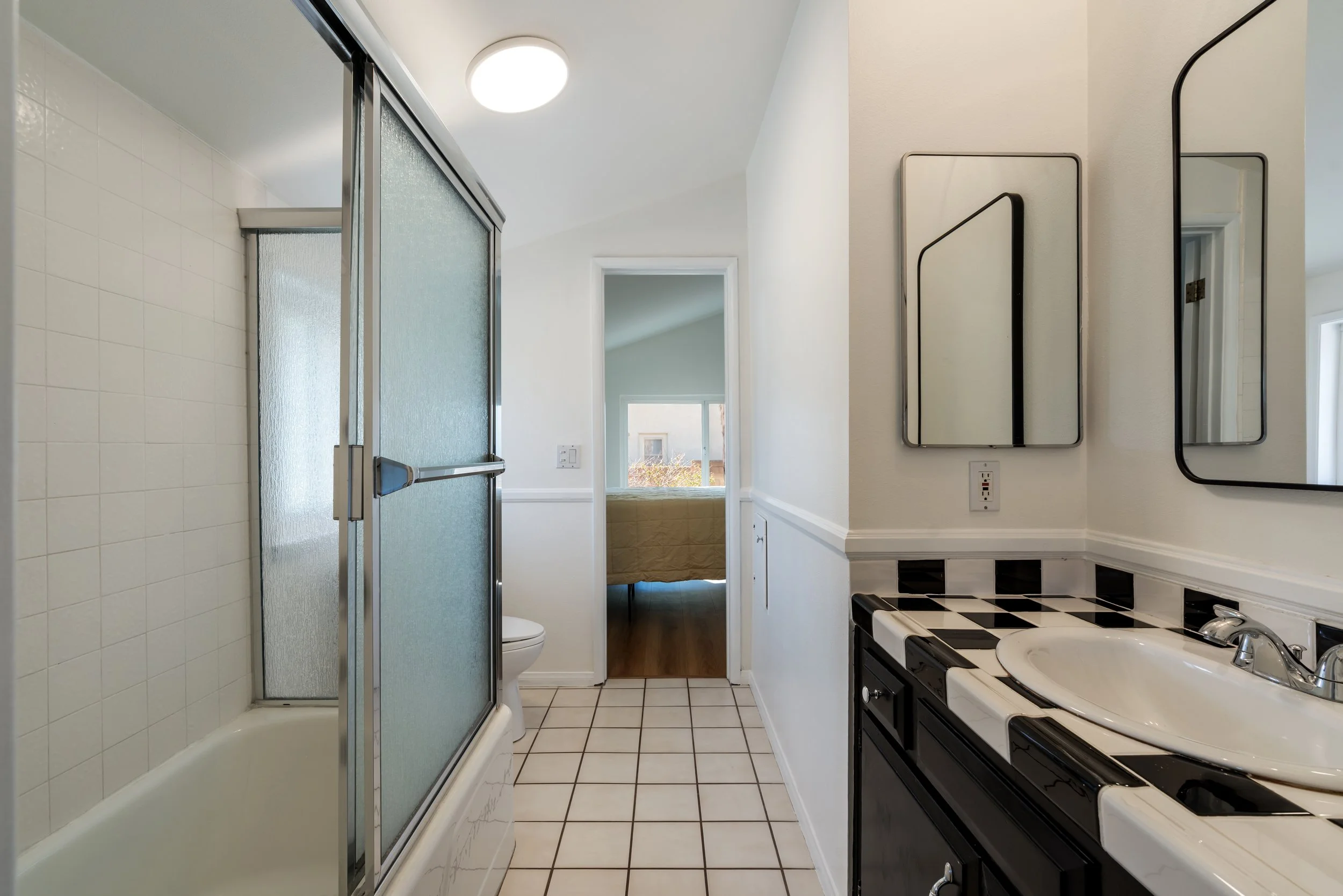 A small bathroom with a bathtub and shower on the left, a toilet next to it, and a vanity with a mirror and sink on the right. There is a window in the adjacent room visible through an open door at the back.