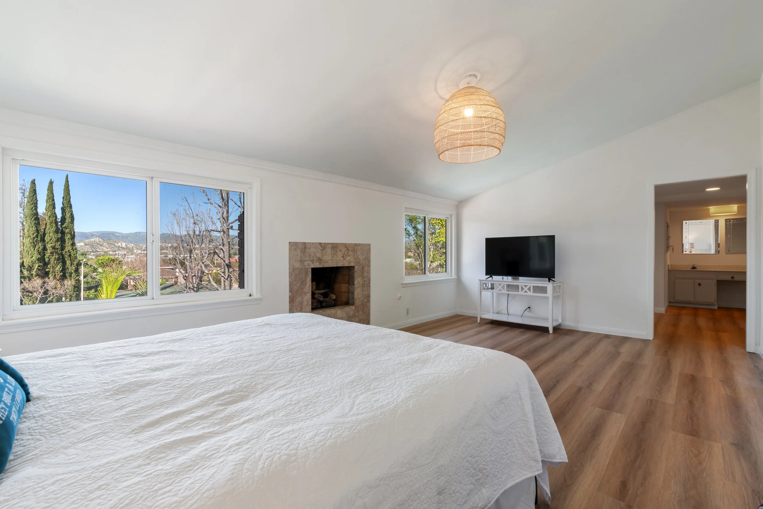 Bright bedroom with large windows, a white bed, a fireplace, a flat-screen TV, and wood flooring.