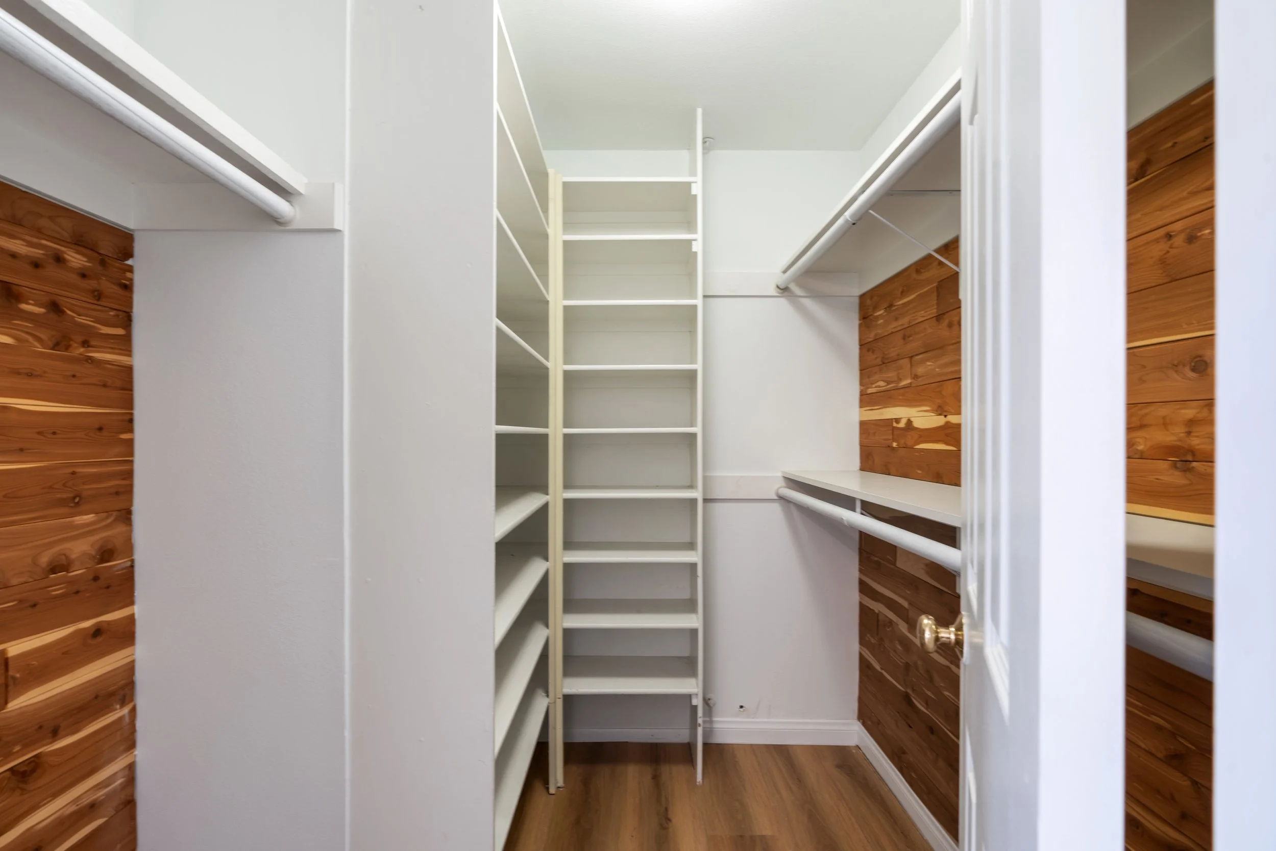 Empty walk-in closet with white shelves, hanging rods, and wood-paneled walls.
