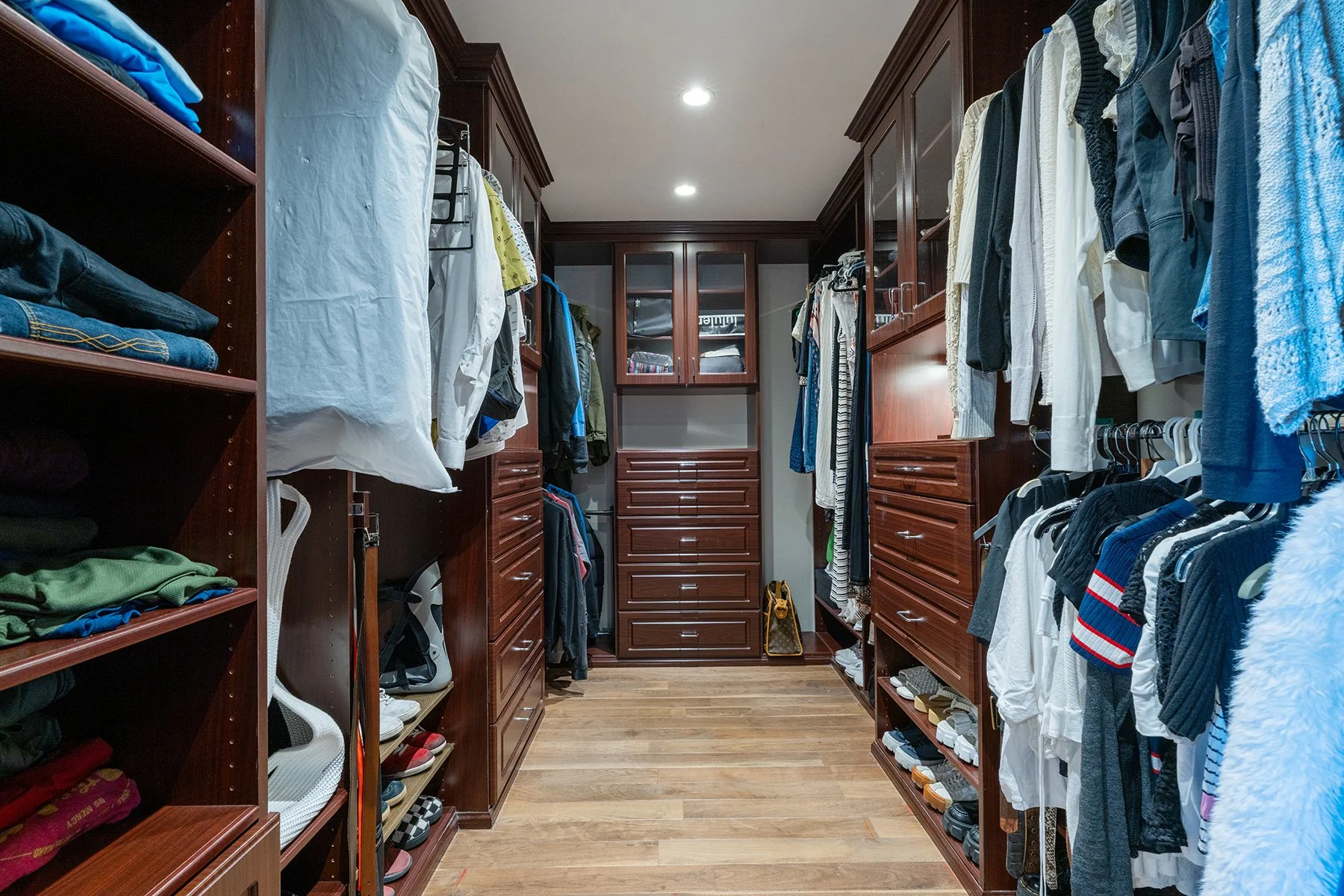 A walk-in closet with built-in wooden shelves and drawers, filled with folded clothes, shoes, and hanging garments arranged on both sides.