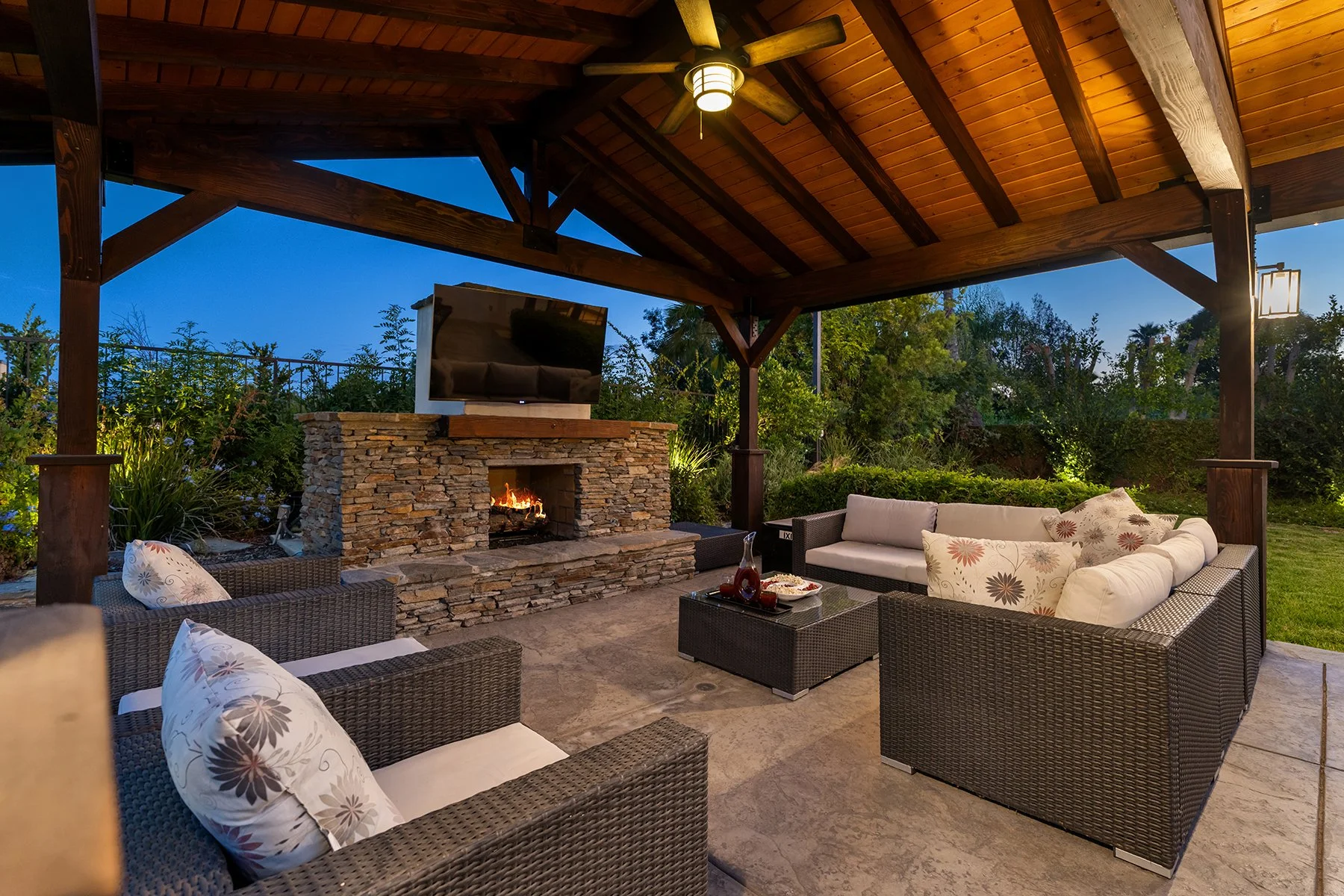 Outdoor patio with wicker seating, cushions, a stone fireplace with a fire, a mounted flat-screen TV, and a ceiling fan under a wooden roof at dusk.