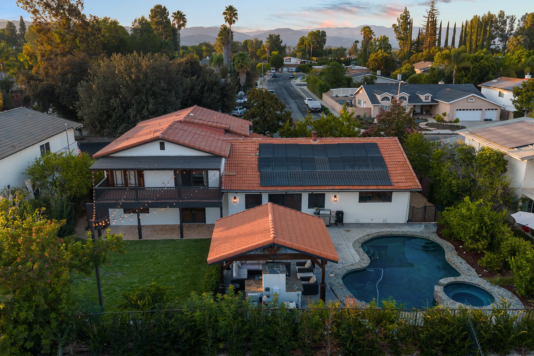 A residential backyard with a swimming pool, hot tub, and a covered outdoor seating area. House has solar panels on the roof. Surrounding trees and neighboring houses visible under a sunset sky.