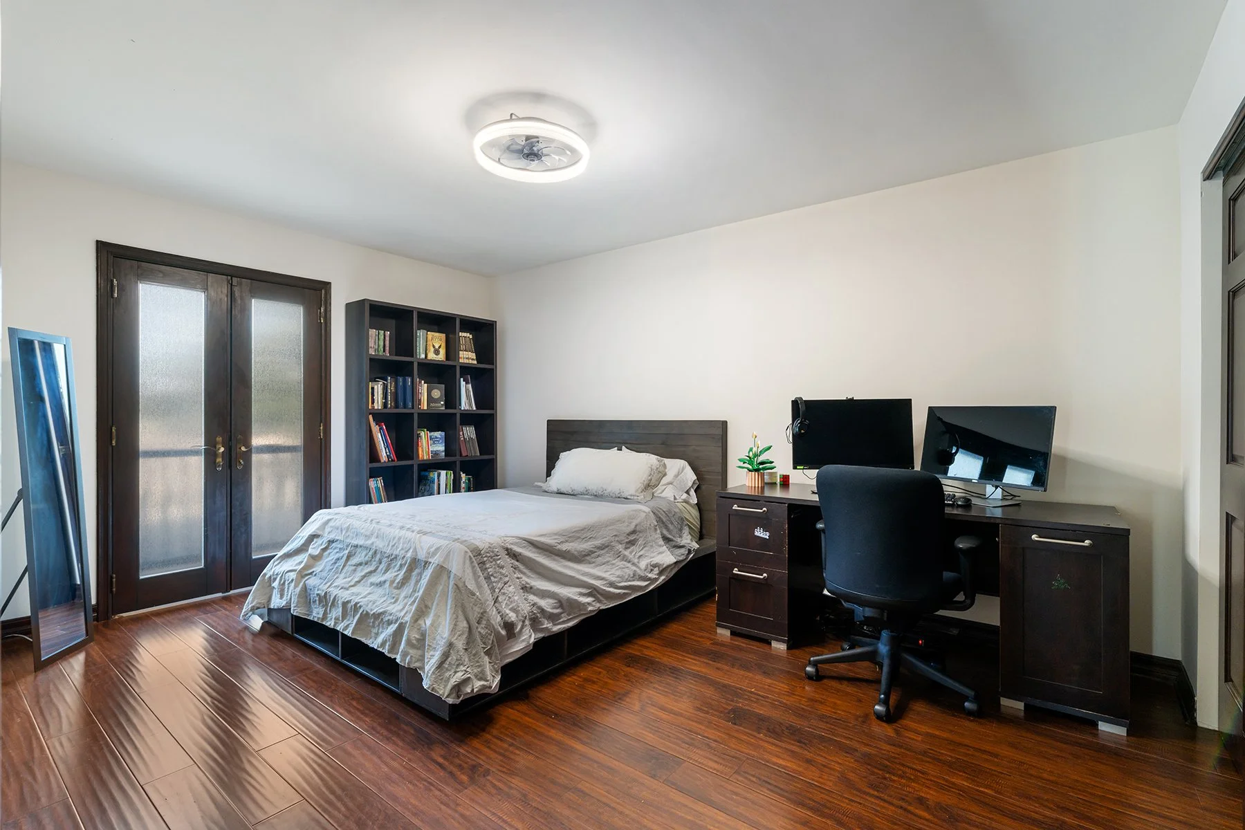 A bedroom with a bed, a black bookshelf filled with books, a double door with frosted glass, a mirror, and a desk with two computer monitors, a chair, and a small potted plant. The room has wooden flooring and white walls.