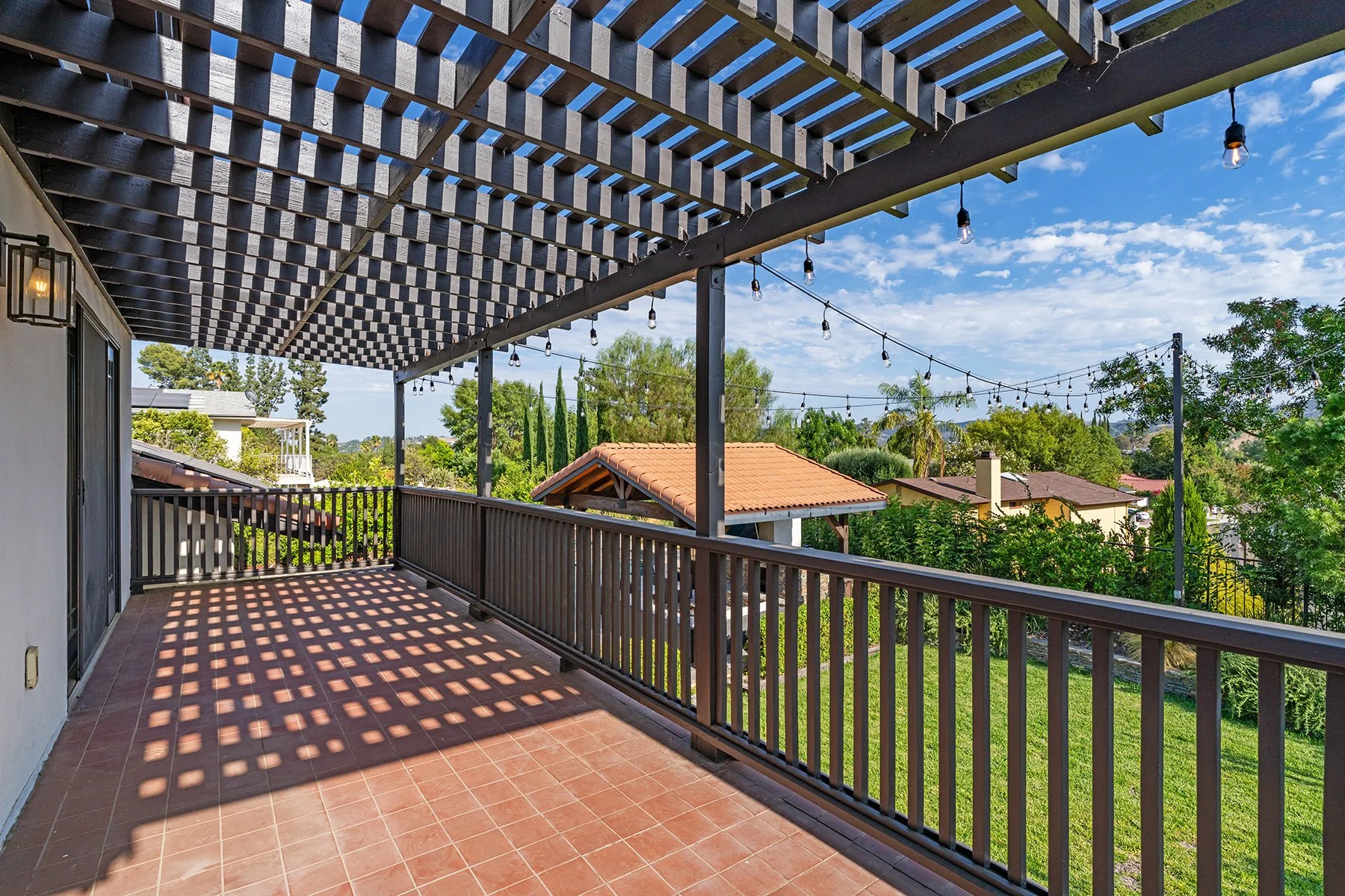 Empty balcony with a wooden lattice overhead casting shadows, string lights hanging, and a view of neighboring houses with tile roofs and surrounding greenery under a partly cloudy sky.