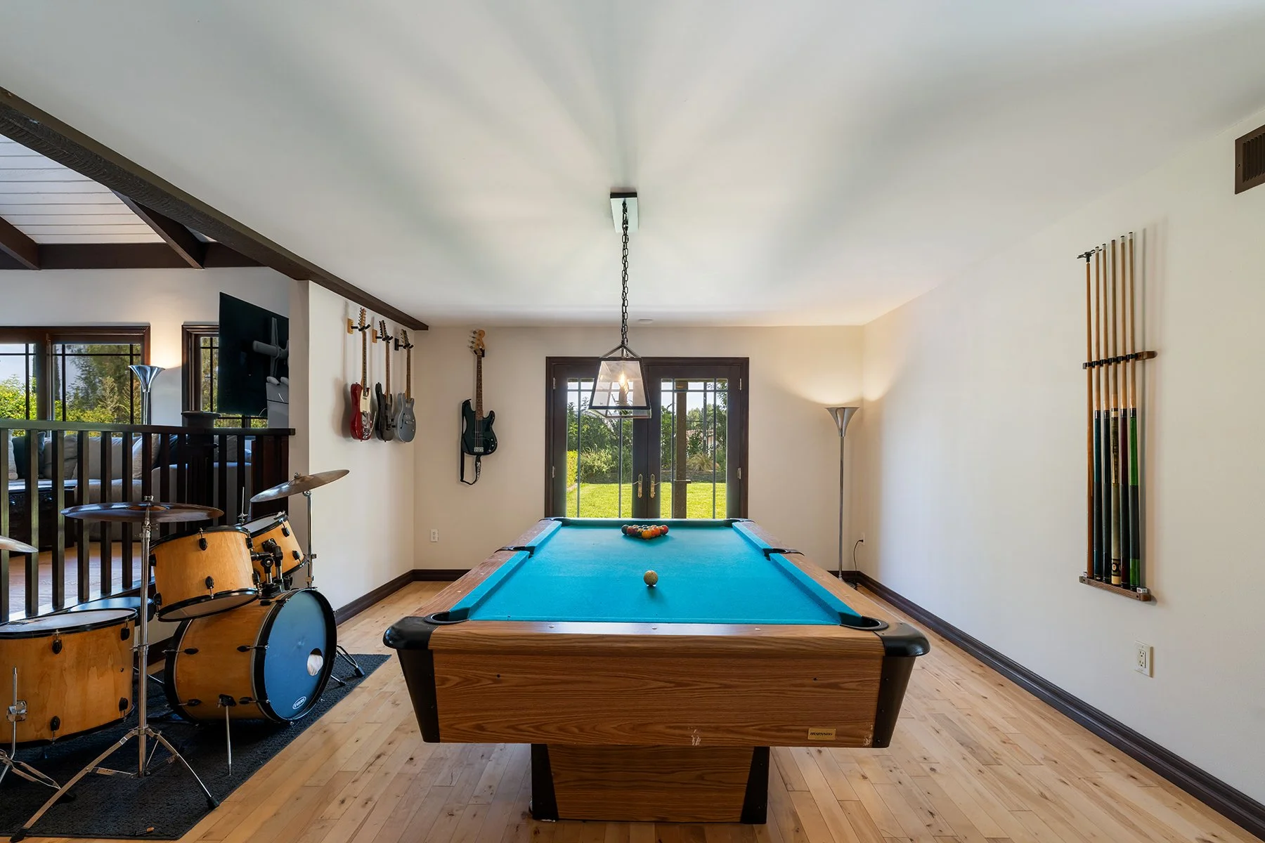 A game room with a pool table, musical instruments on the wall including guitars and drums, and a view of a backyard through glass doors.