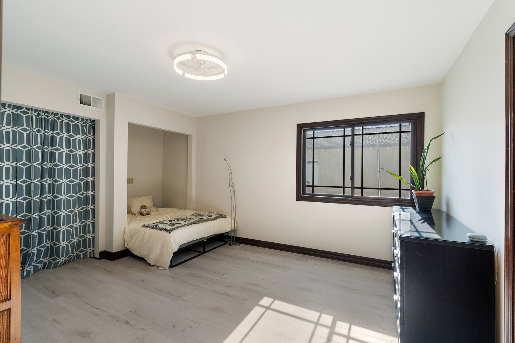A minimalist bedroom with a single bed, a black dresser with a potted plant, a window with dark wood framing, and a ceiling light fixture.