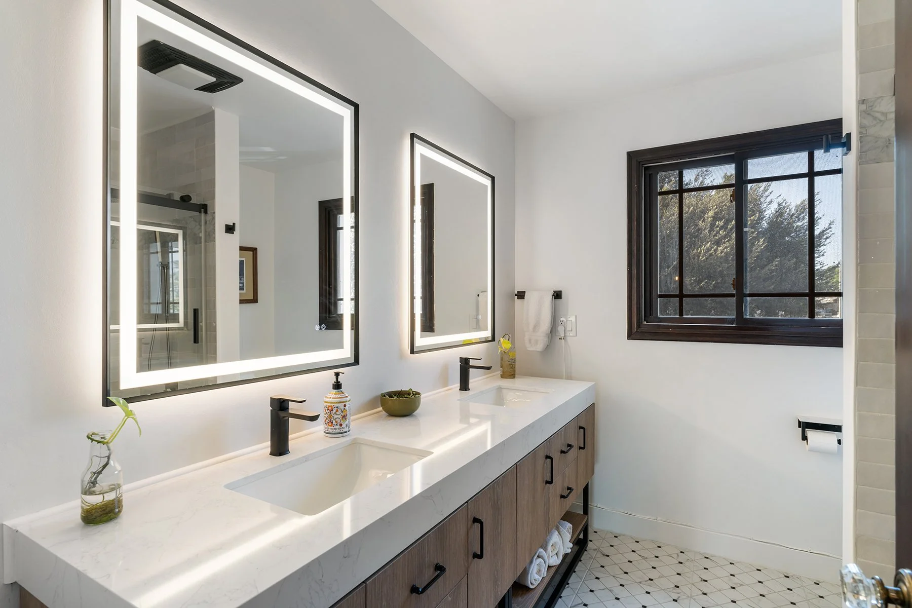 Modern bathroom with double sinks, illuminated mirrors, wooden cabinetry, and a window showing trees outside.