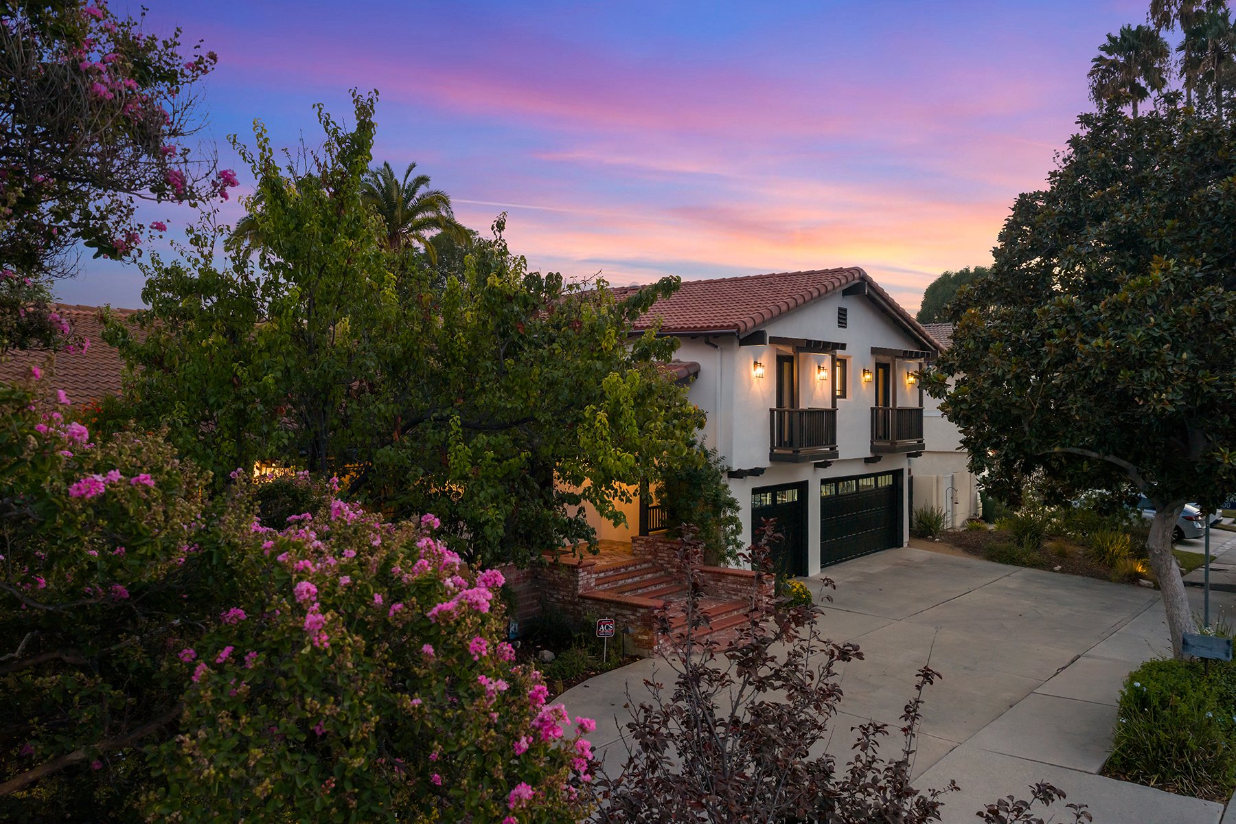 A two-story house with a red tile roof, white walls, and black window frames, surrounded by lush greenery and pink flowering bushes, under a colorful sunset sky with pink, purple, and blue hues. Represented by Phil Missig and the Missig Team.