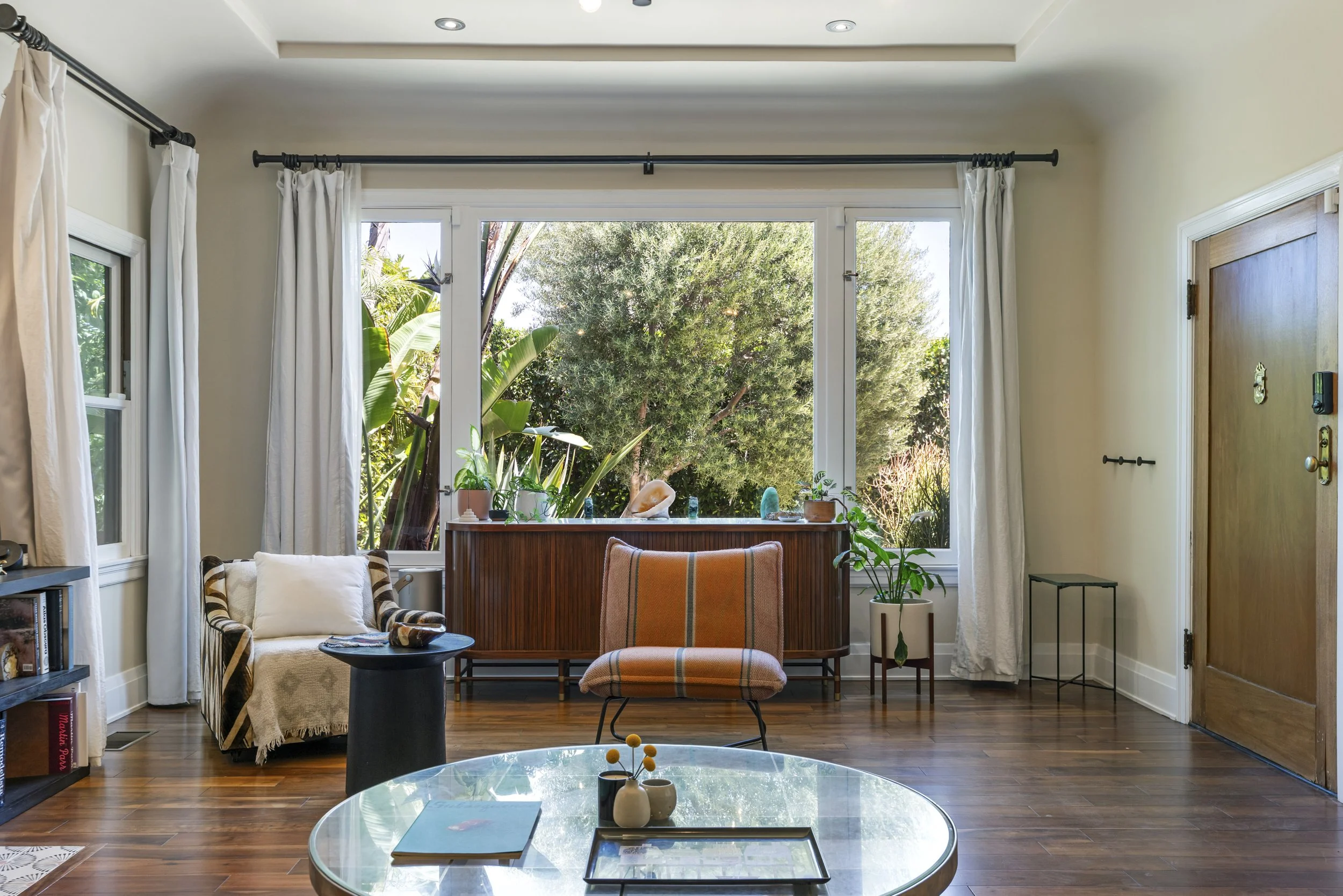 Living room with a large window overlooking greenery, beige curtains, a plaid armchair with a blanket, a small round black table, potted plants, and bookshelves.