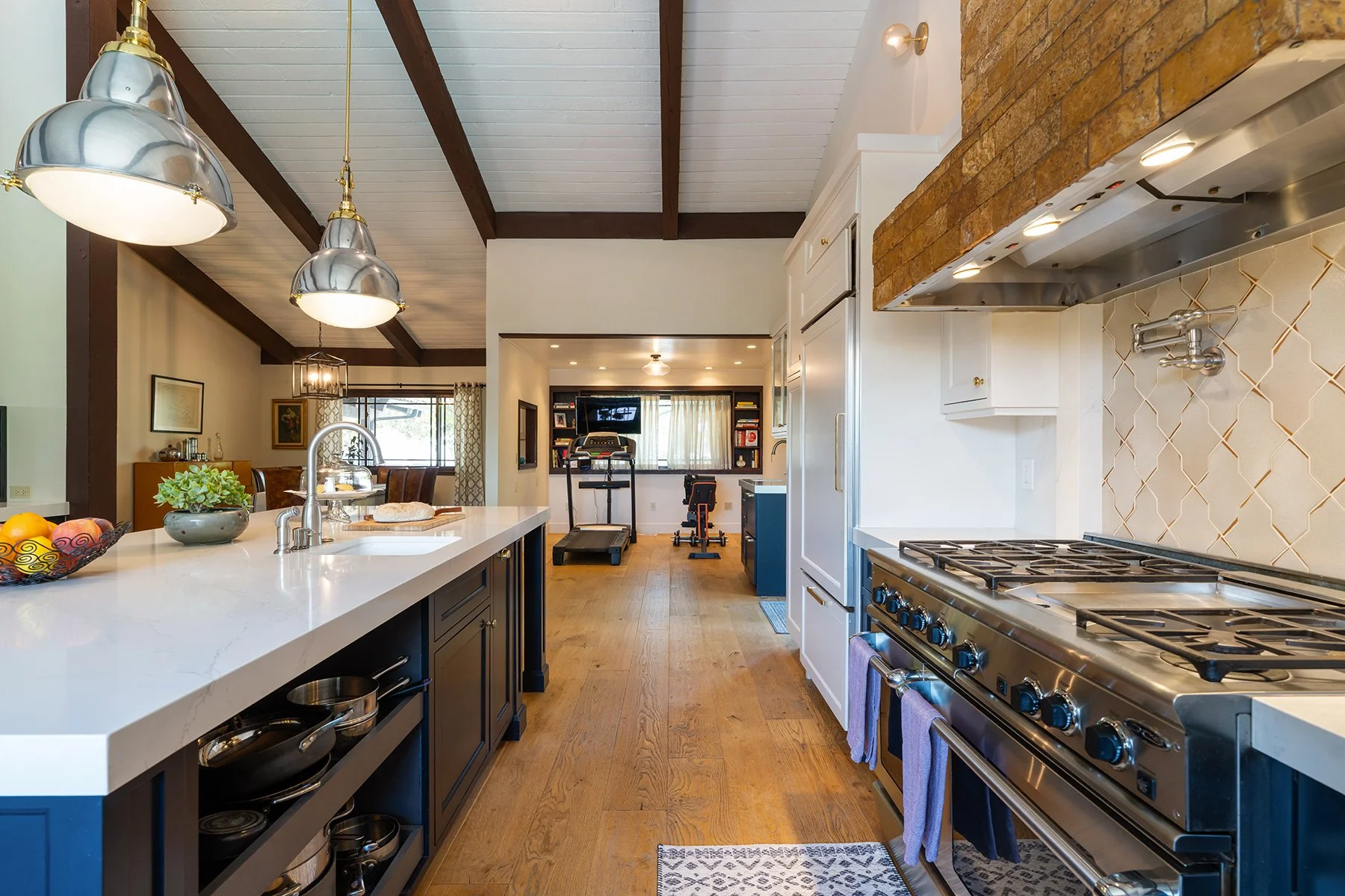 Interior view of a modern kitchen with a white island countertop, black lower cabinets, stainless steel oven, and a brick range hood. The space opens to a living area with gym equipment and a television. Wood flooring and exposed ceiling beams add warmth to the room.