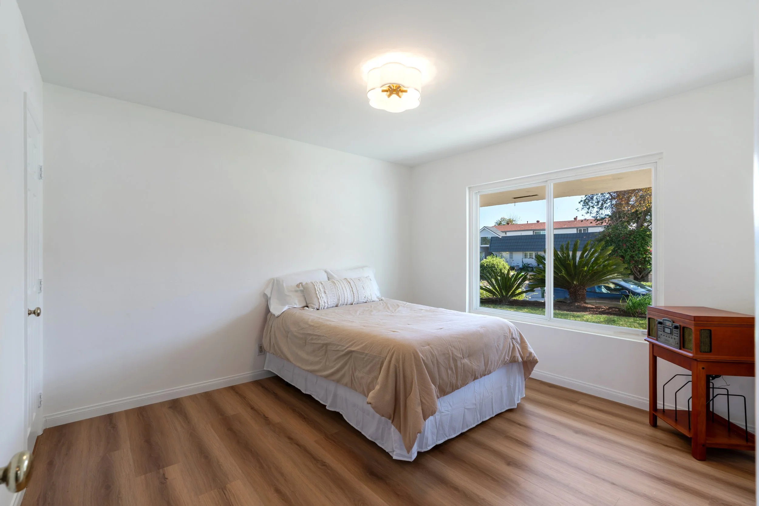 A bedroom with a bed, nightstand, large window showing palm trees outside, wood flooring, and white walls.