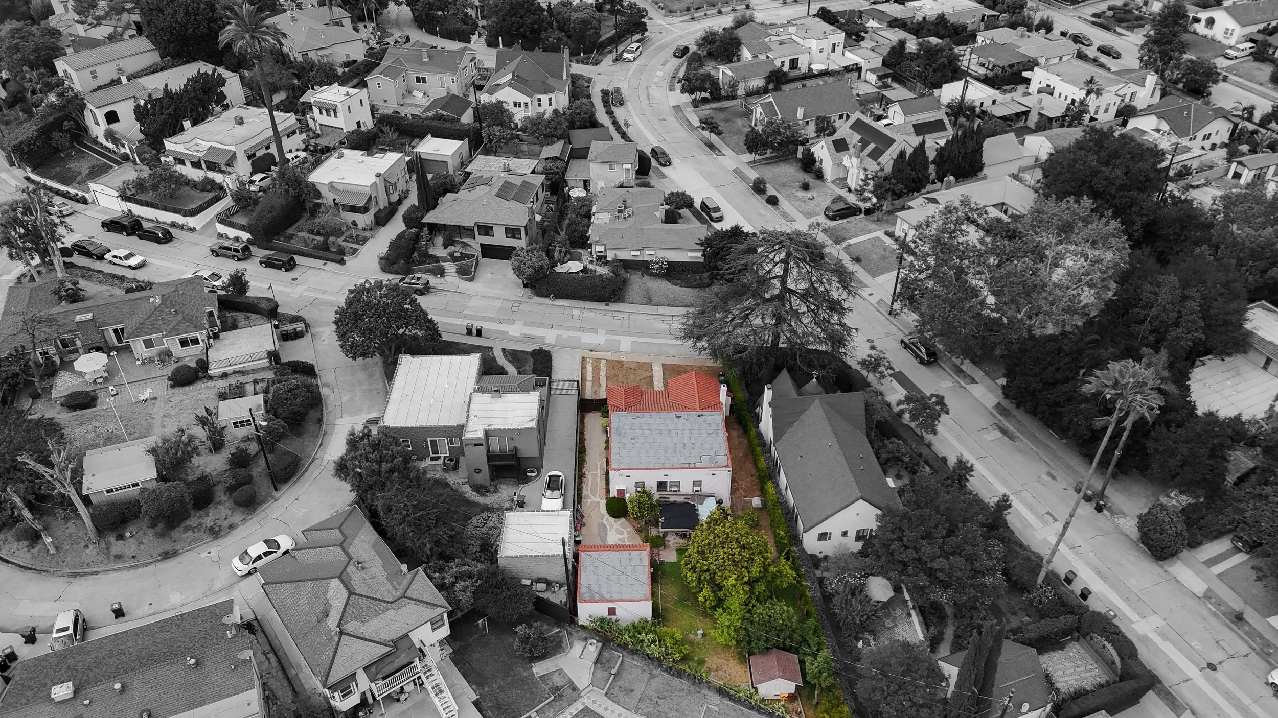 Aerial view of a suburban neighborhood with houses, trees, and cars on the streets. One house outlined in color stands out among the black and white surroundings.