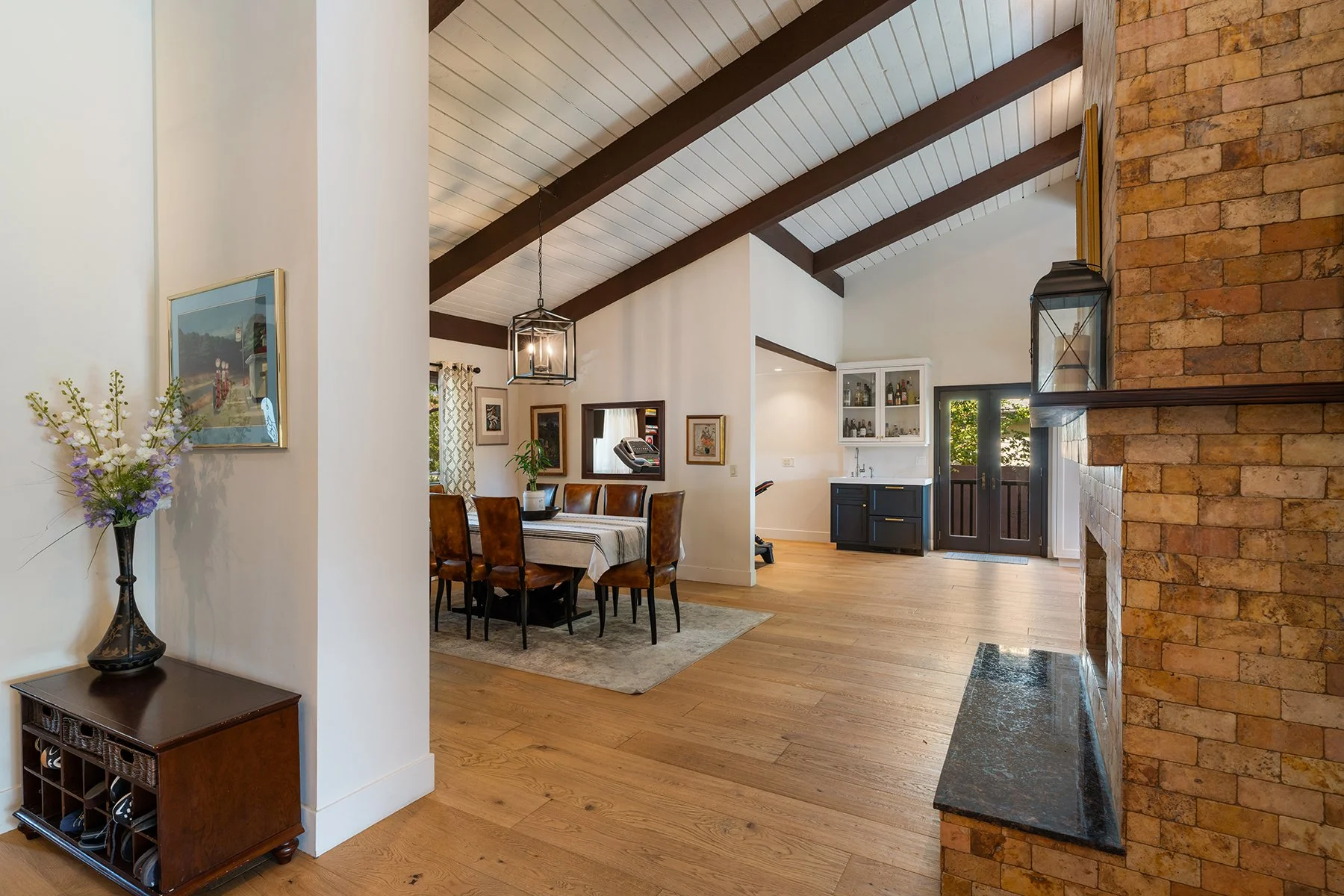 Interior view of a living and dining area with wooden flooring, white walls, and a vaulted ceiling with dark wooden beams. There is a dining table with six chairs, a corner kitchen with black cabinets, and a brick fireplace with a black marble mantle.