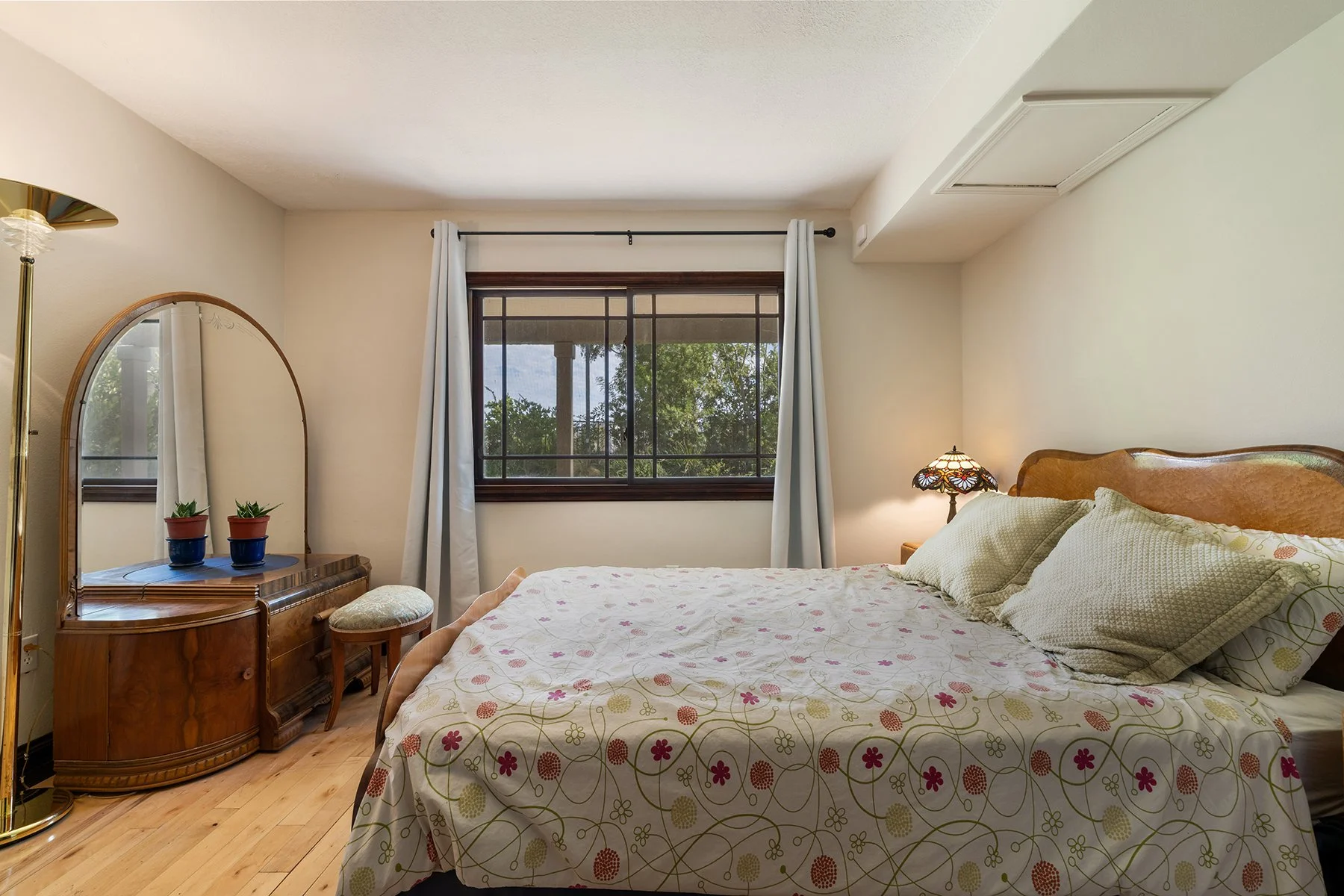 A cozy bedroom with a large bed covered in a floral patterned quilt, two pillows, and a wooden headboard. There is a dresser with a mirror and two small potted plants on top, and a cushioned stool next to it. A window with white curtains shows trees outside. A Tiffany-style lamp provides warm lighting on a nightstand beside the bed.