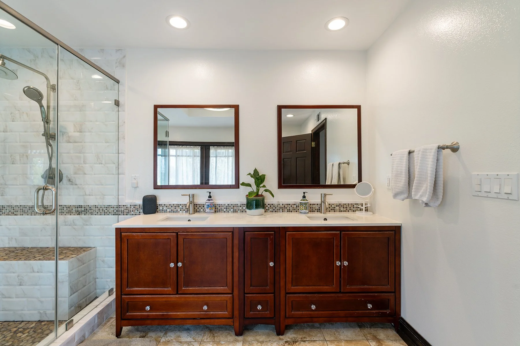 Bathroom with double vanity, wooden cabinets, two mirrors, towels on towel bar, potted plant, soap dispensers, and a walk-in shower with marble tiles and pebble floor.
