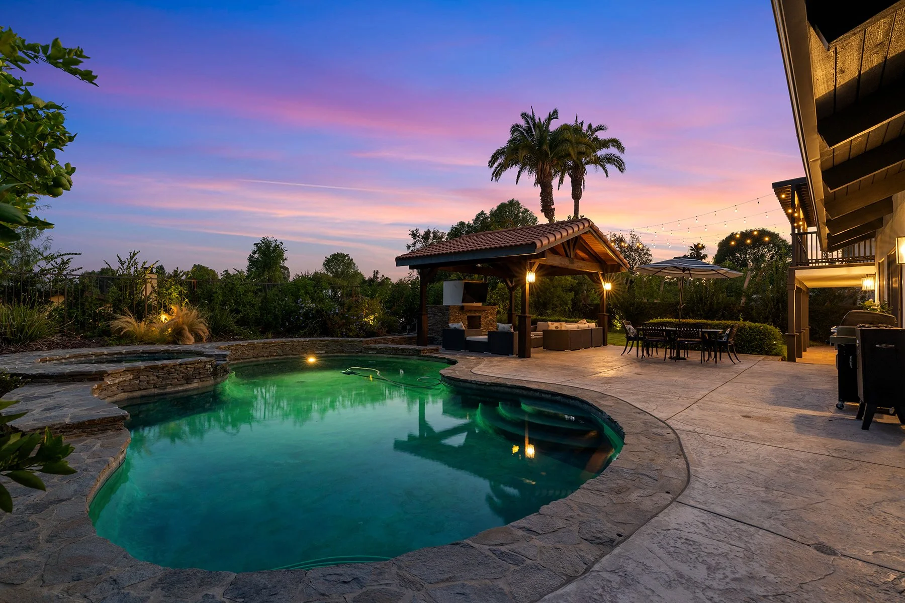A backyard pool area during sunset with colorful sky, palm trees, outdoor seating, a hot tub, and string lights.