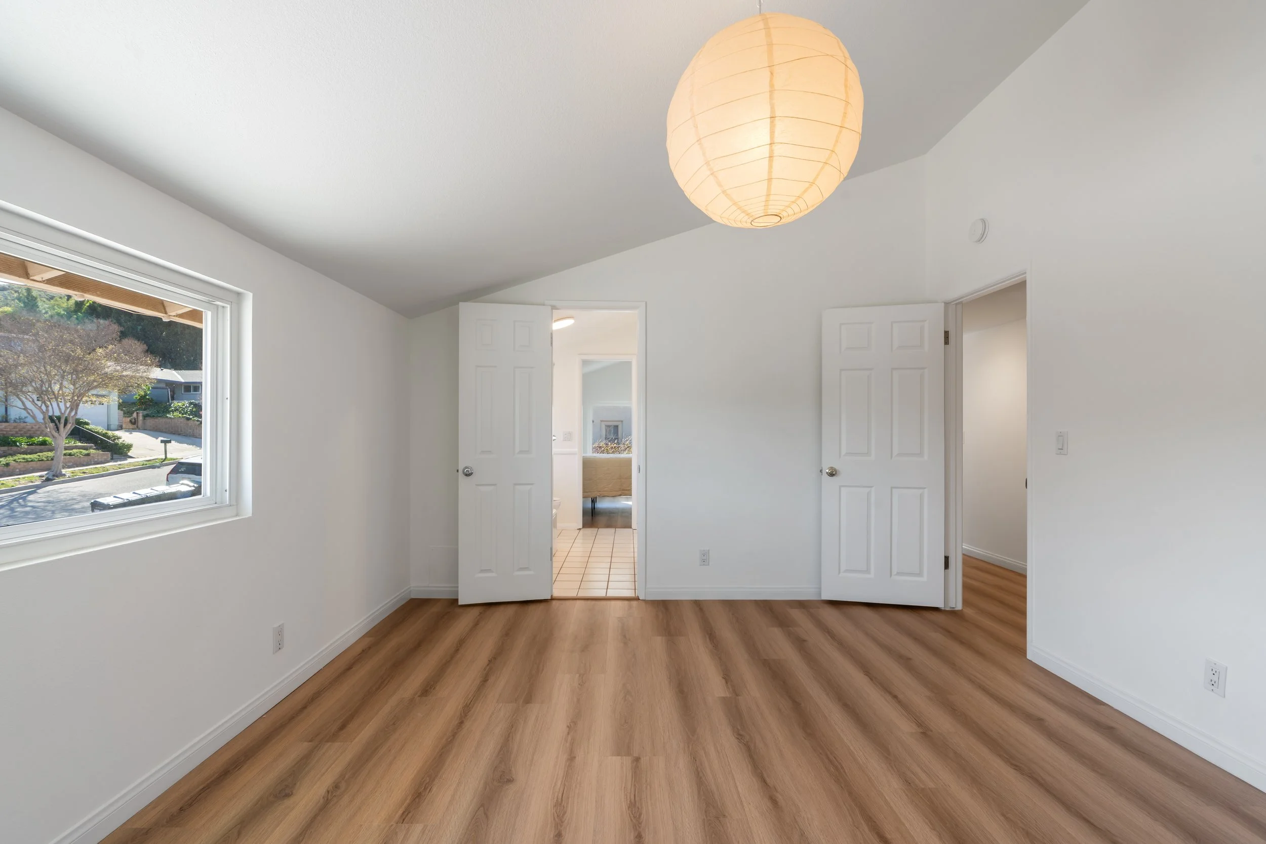 Empty room with light-colored walls, hardwood floor, a window showing a tree and street outside, and two open doors leading to a bathroom and another room, with a paper lantern ceiling light.