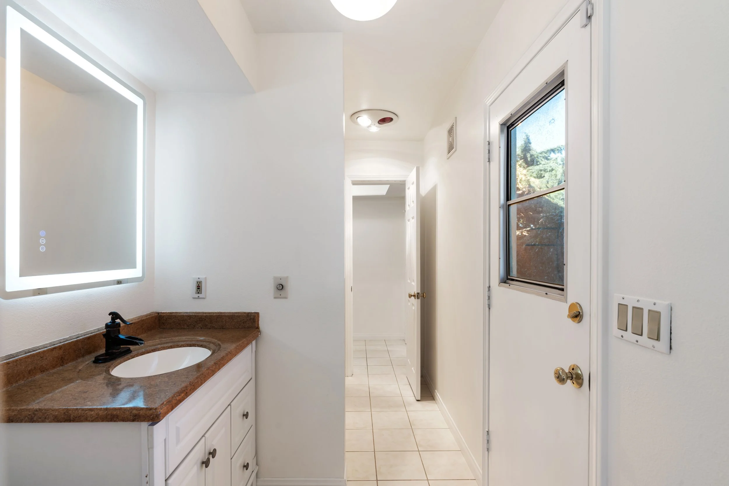 Interior view of a small bathroom with a granite countertop, white cabinets, a rectangular mirror with LED lighting, a black faucet, a window showing outdoor trees, and a door leading outside, with tiled flooring.