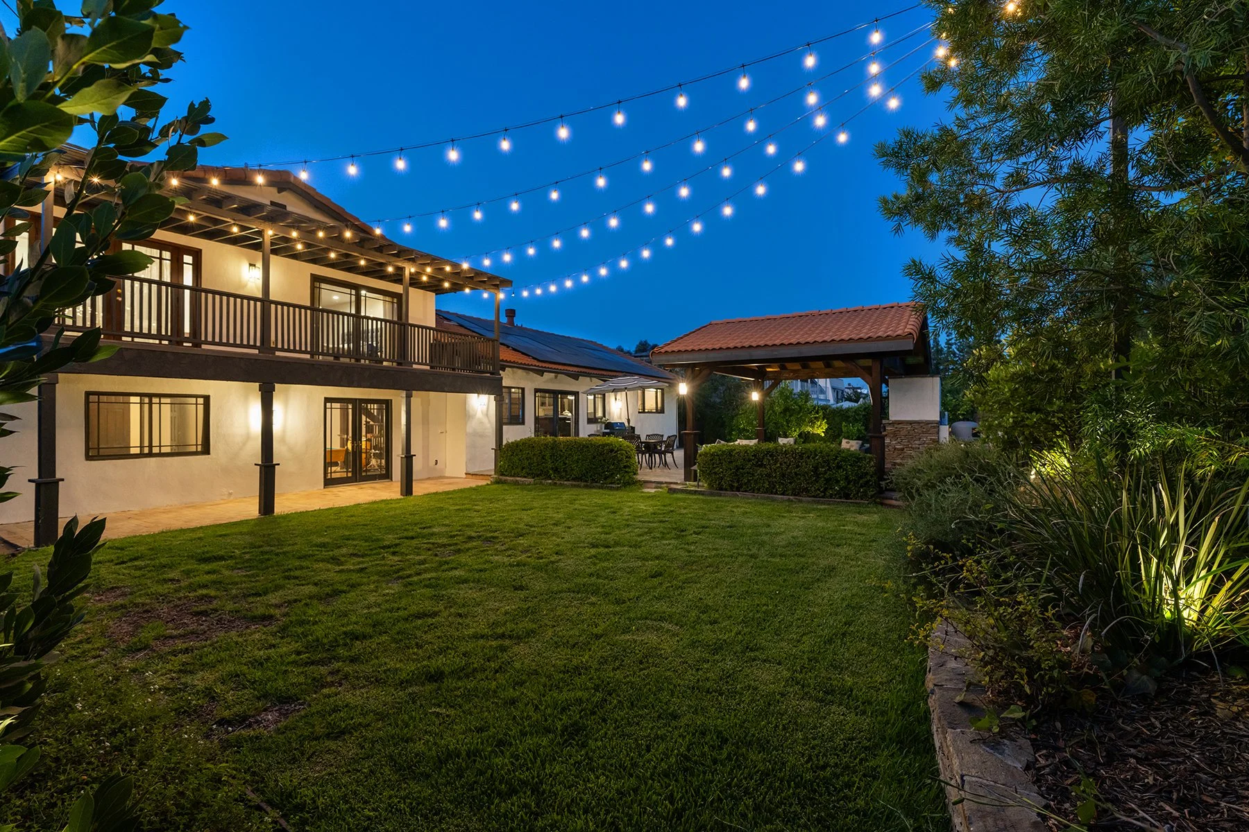A backyard at dusk with string lights, a lawn, a shaded patio area, and a two-story house with windows and a balcony.