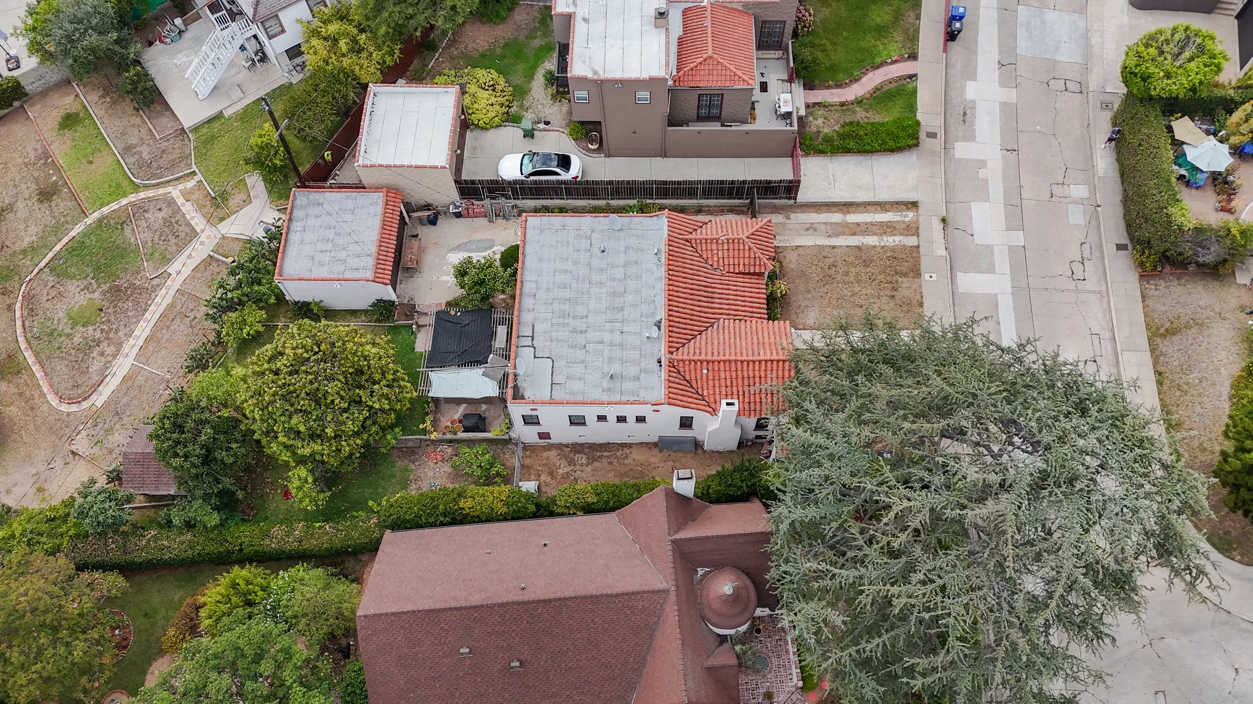 An aerial view of residential houses with trees, a car parked in a driveway, and a backyard with garden beds and pathways.