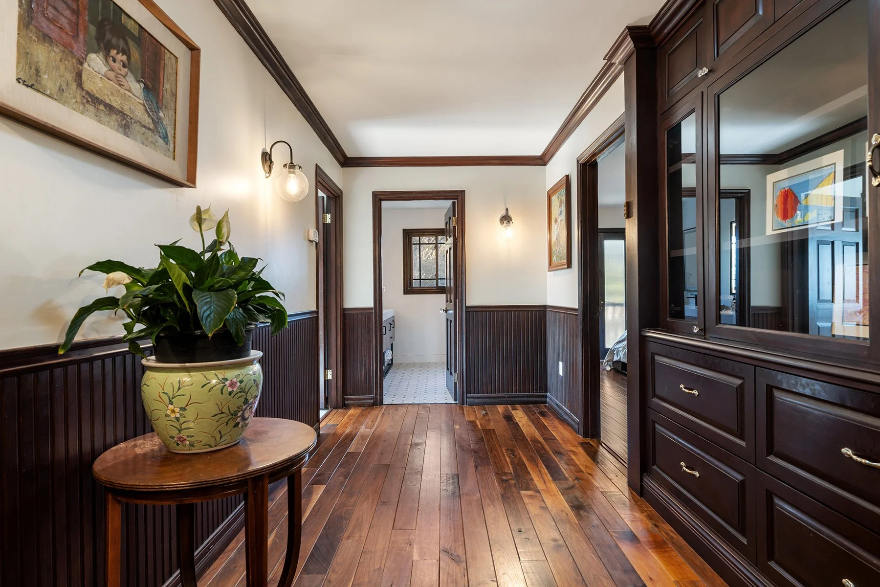 Hallway with wood-paneled walls, hardwood floor, framed artwork, a potted plant on a table, and lights on the wall.