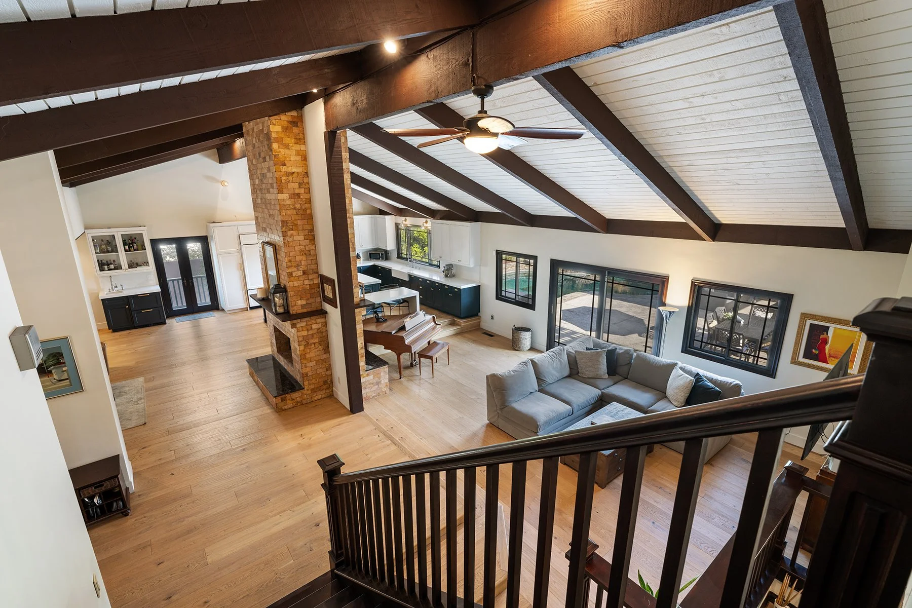 A spacious open living area with a vaulted ceiling supported by exposed wooden beams, a ceiling fan, a brick fireplace, a large gray sectional sofa, a hymn piano, and multiple windows and glass doors leading outside.