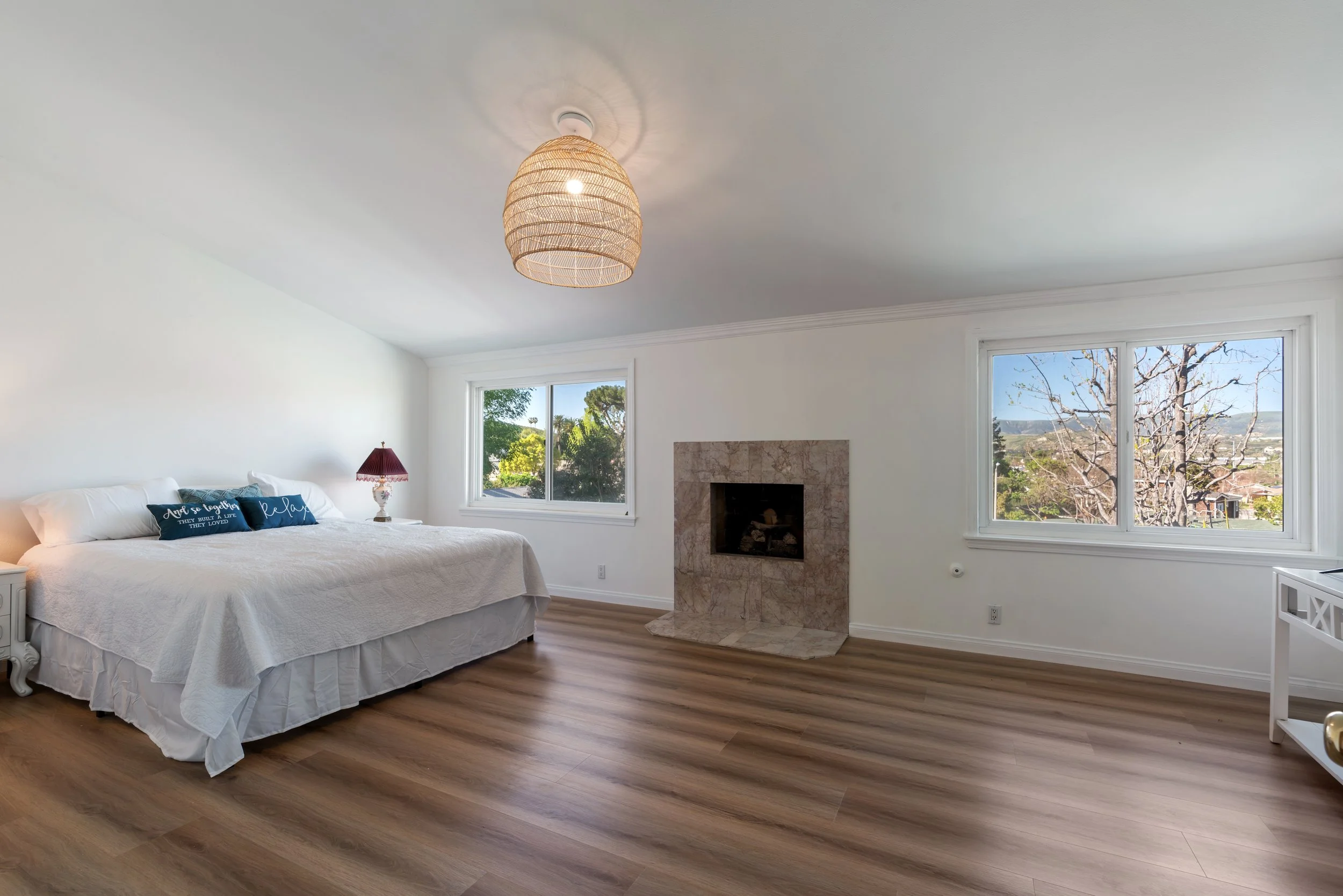 A bright, spacious bedroom with a white bed, a maroon bedside lamp, a stone fireplace, and large windows showing trees and hills outside.