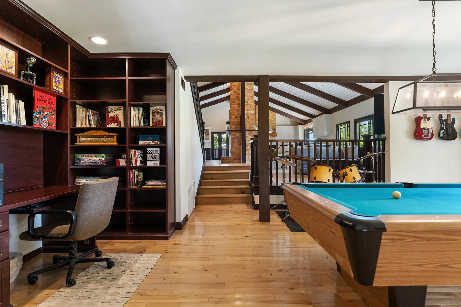 Interior of a recreation or game room with a pool table, electric drum set, wall-mounted guitars, a bookshelf, a desk with a chair, and a staircase leading to an upper level with wooden beams and brick chimney.