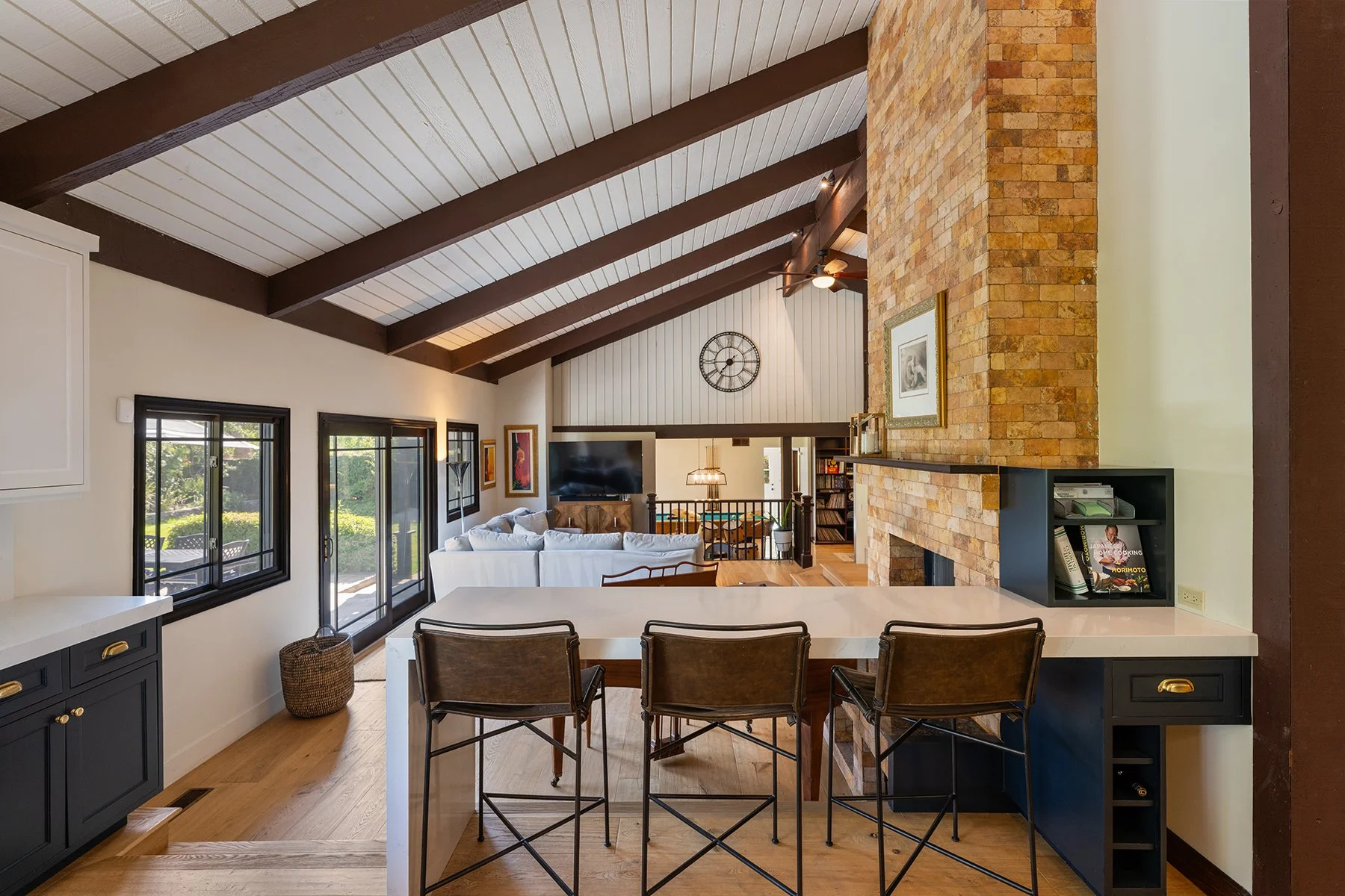 Interior view of a living space seen from the kitchen area with barstools, a white sofa, a brick fireplace, large windows, and decorative wall art, with a high ceiling featuring dark wooden beams