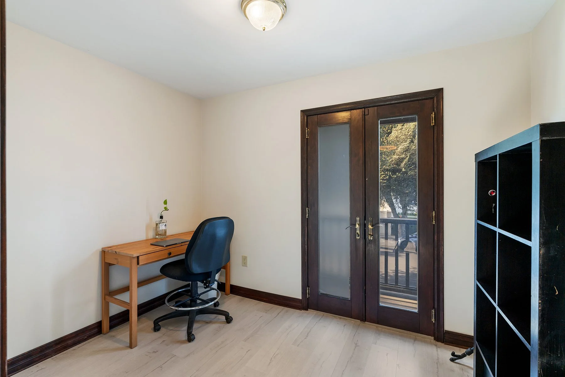 Small home office with a wooden desk, black chair, and a small plant in a jar. Double glass doors lead outside, with trees visible. Black bookshelf on the right side.