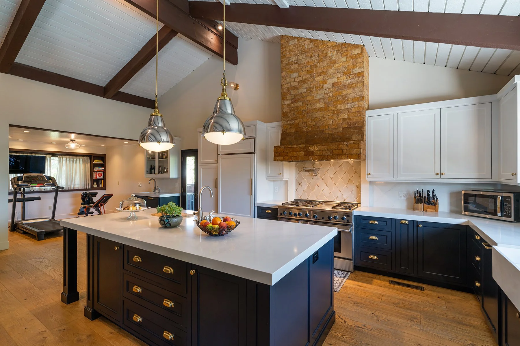 Modern kitchen with black and white cabinets, a large white island with black base, a brick chimney, stainless steel appliances, pendant lighting, and a view into a living area with treadmill and exercise equipment.