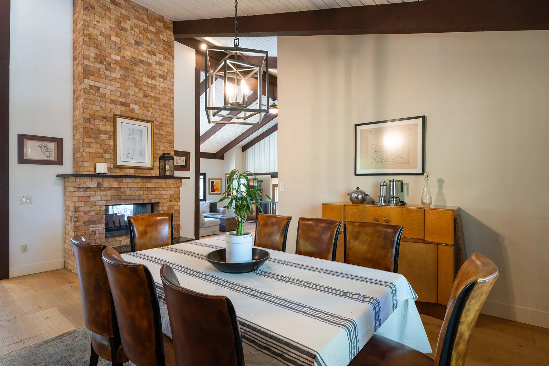 Interior of a dining room with a brick fireplace, a wooden sideboard, and a rectangular table with a striped tablecloth and chairs. There is a potted plant on the table, artwork on the walls, and modern lighting fixtures.