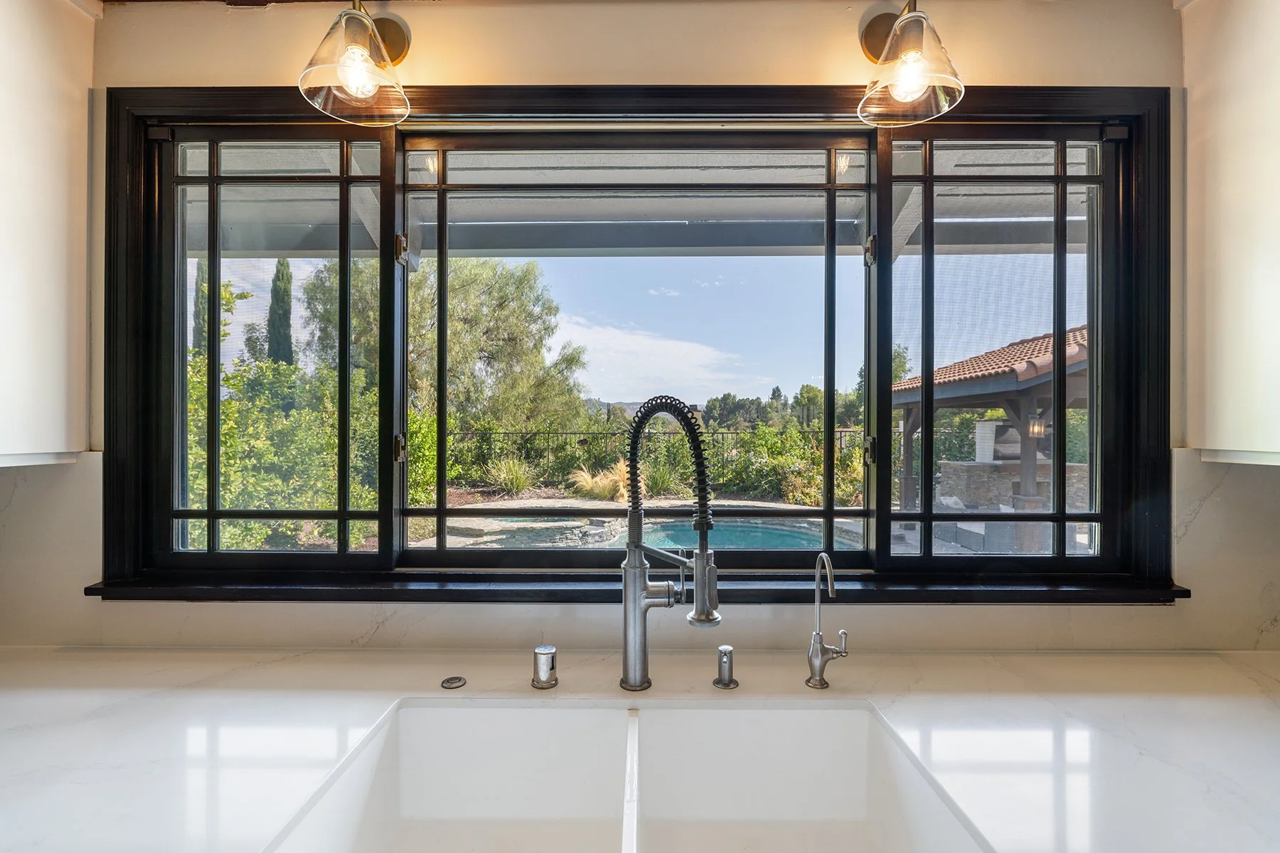 View through a large kitchen window showing a backyard with trees, bushes, and a patio with a pergola and a pool, with a white kitchen sink and faucet in the foreground.