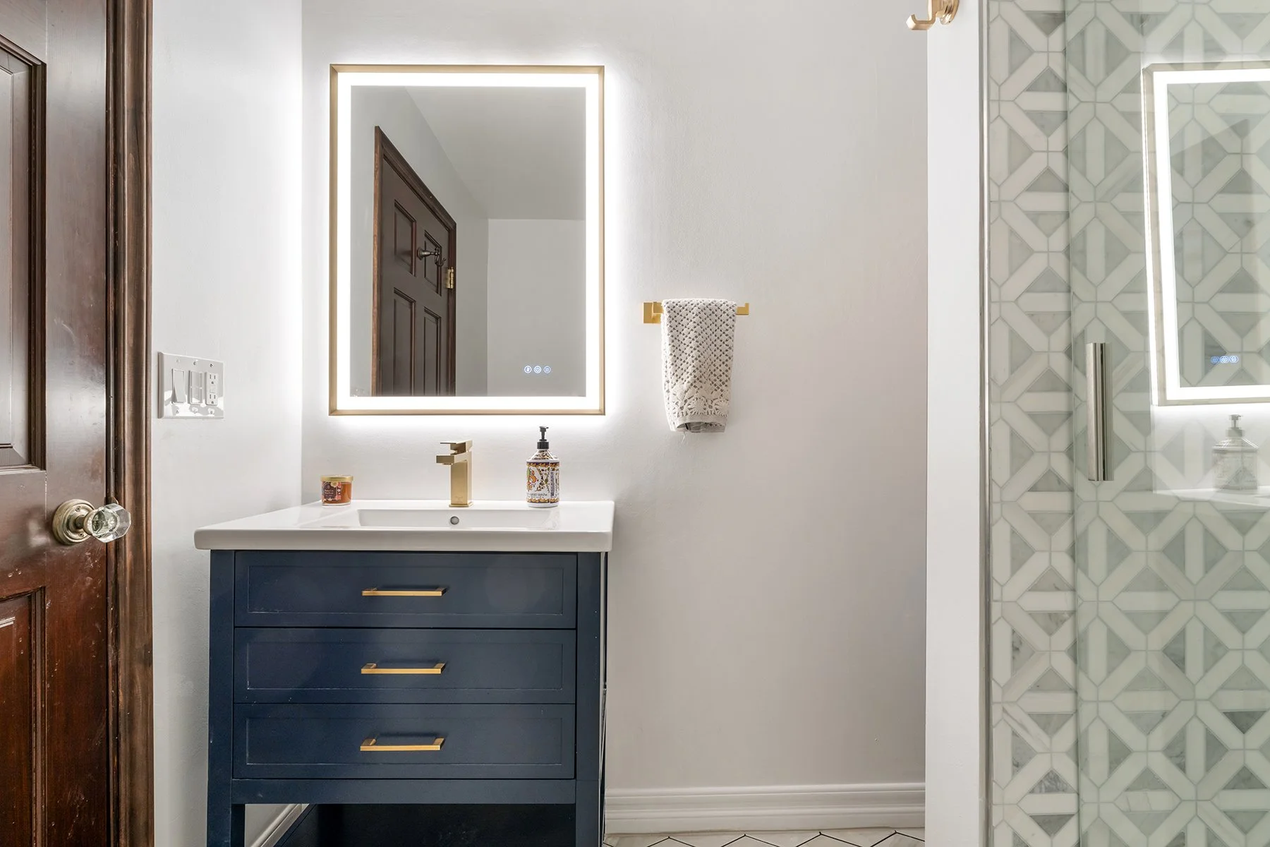 A modern bathroom with a navy vanity, gold handles, and a white countertop with a faucet. Above the sink is a backlit mirror. To the right, a towel hangs on a gold towel bar, and a patterned soap dispenser is on the counter. Part of a glass-enclosed shower with geometric patterned tiles is visible on the right, and a wooden door with a brass doorknob is on the left.