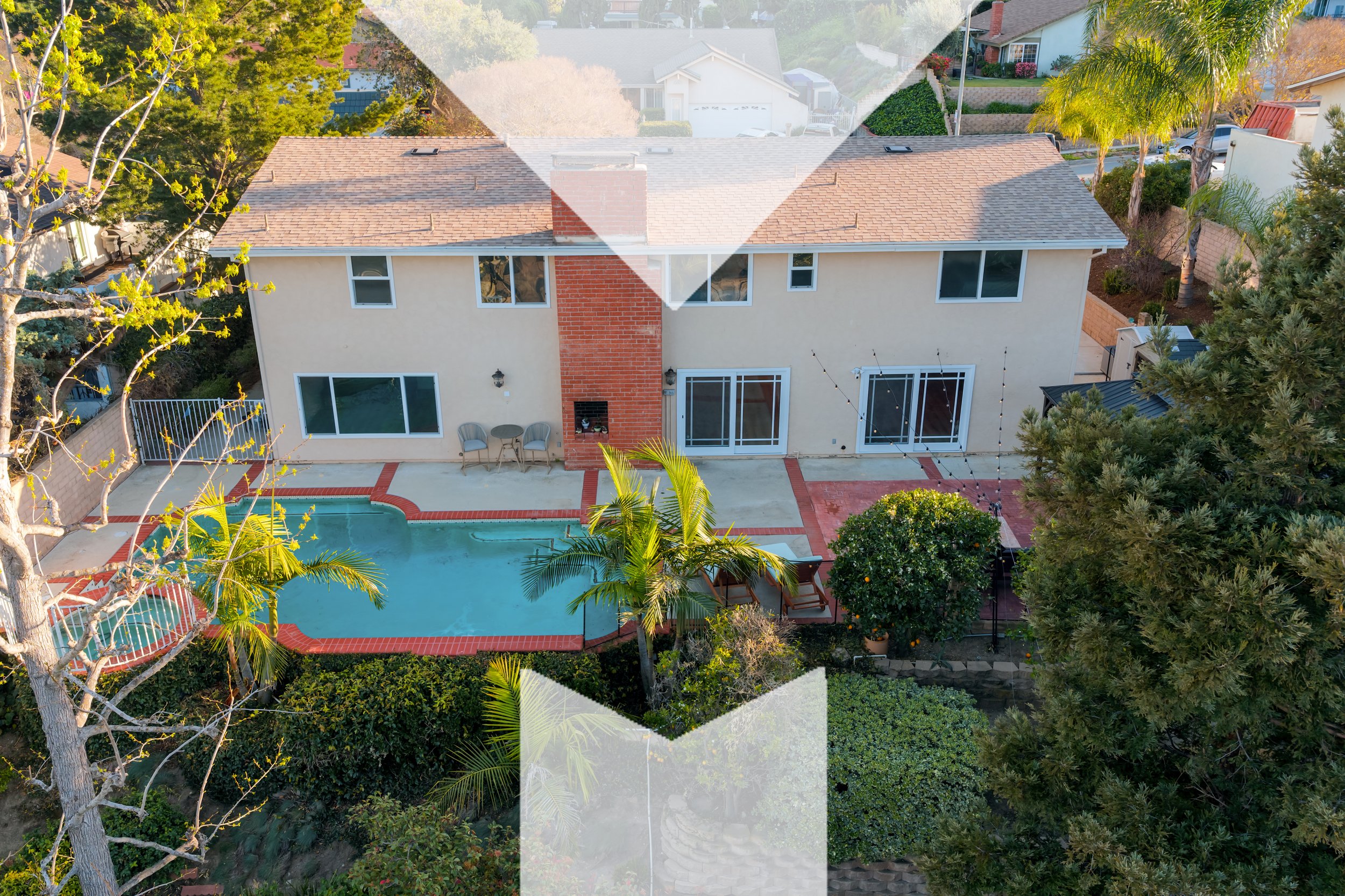 Aerial view of a backyard with a swimming pool, palm trees, and patio furniture, adjacent to a beige two-story house with large windows and a brick chimney, surrounded by trees and neighboring houses.