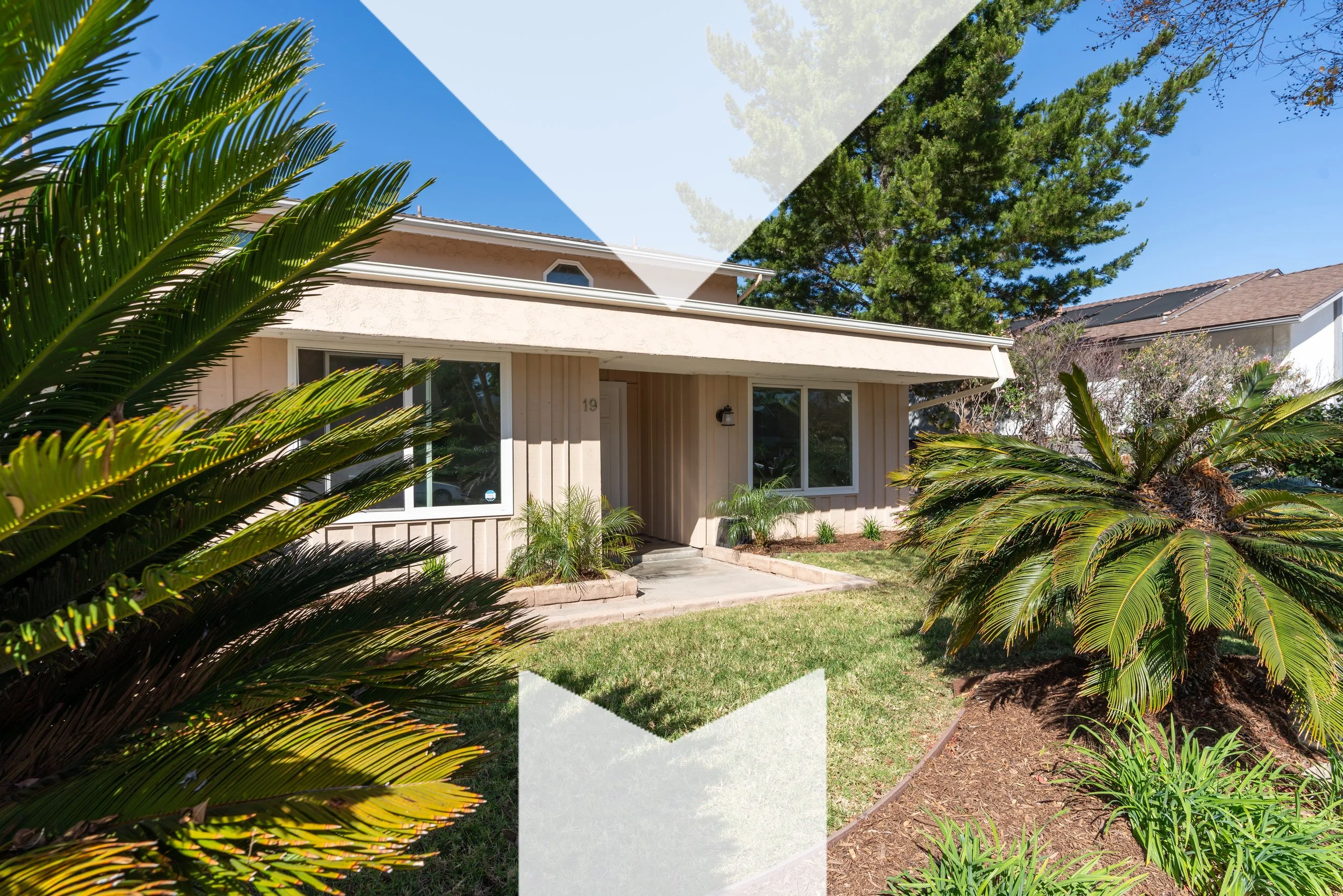 Front view of a beige house with large windows, a small porch, surrounded by lush palm plants and a well-maintained lawn, under a clear blue sky.