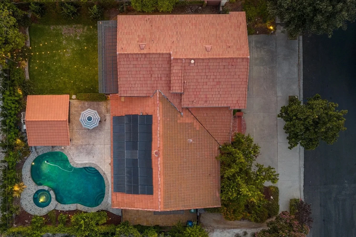 Aerial view of a house with a red tile roof, solar panels on the roof, a backyard with a swimming pool, a patio, and yard decorations, surrounded by trees and greenery.