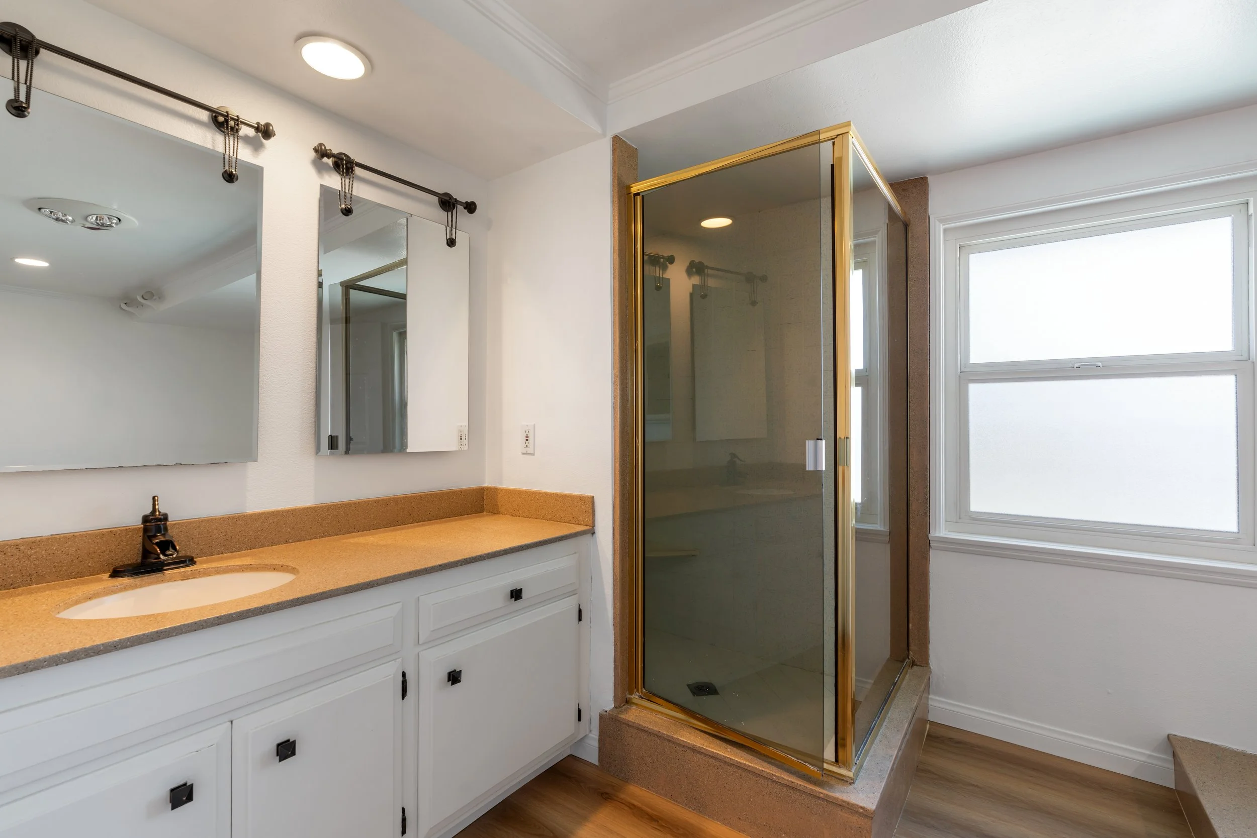 A bathroom with a white vanity and large mirror, a glass shower enclosure with a gold frame, and a double-pane window with frosted glass.
