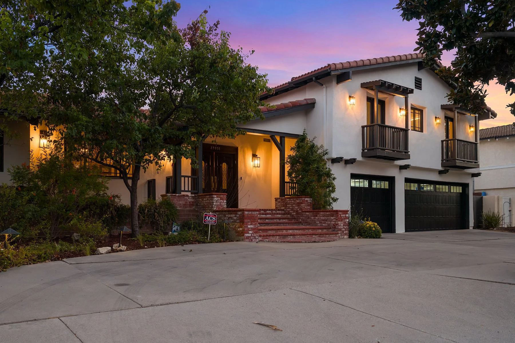 A two-story house with a white exterior, red tile roof, and dark wooden accents, including balconies and window frames. The house has exterior lighting on, illuminating the entrance and upper level. In front, there are brick steps leading to the front door, surrounded by landscaped plants and a driveway.