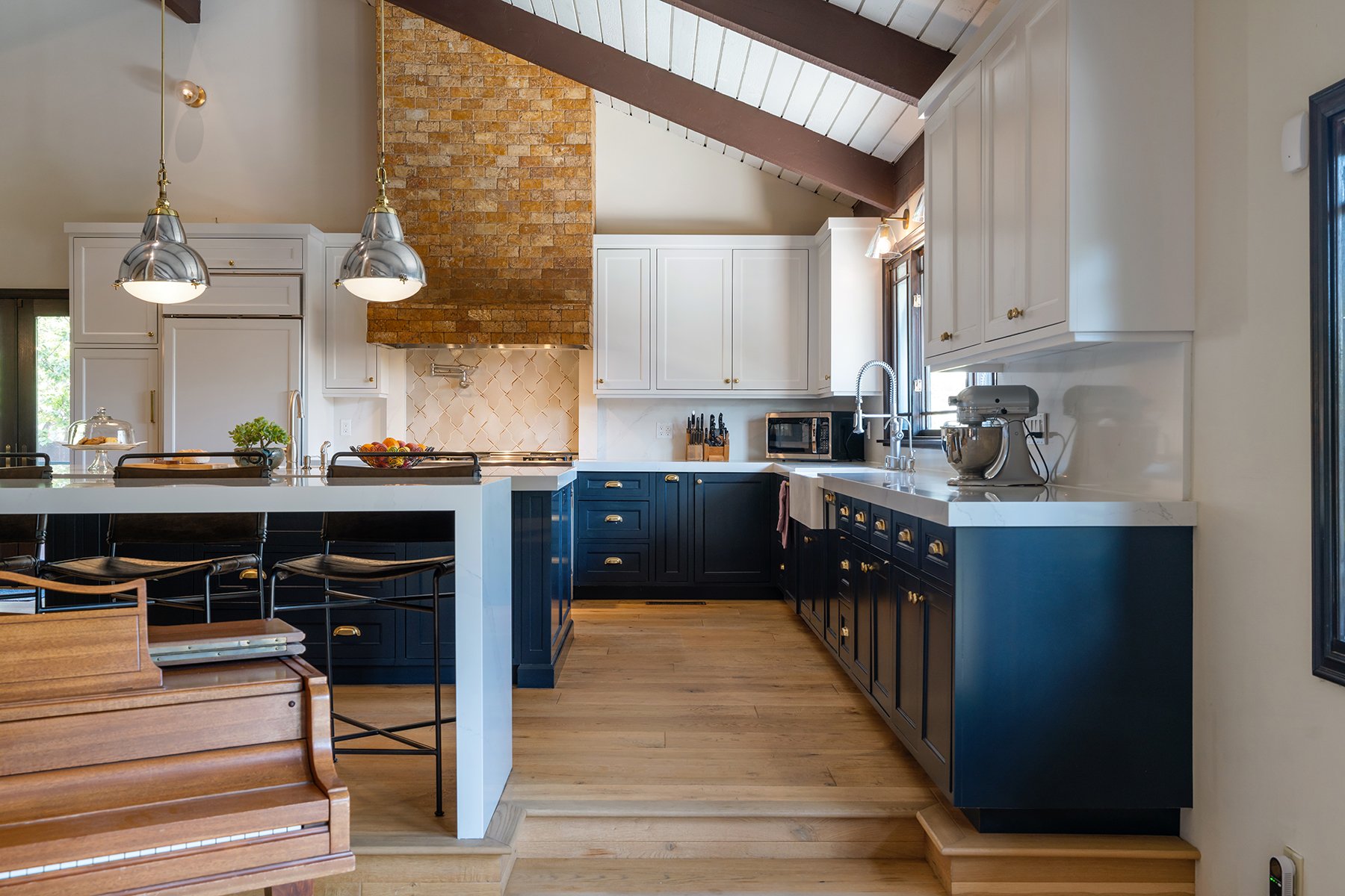 Modern kitchen with white and navy blue cabinets, brick chimney, wood flooring, and stainless steel appliances.