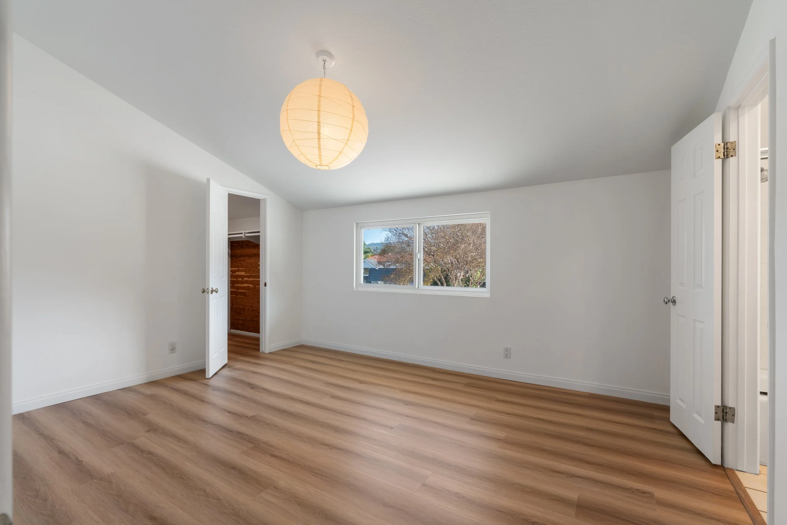 Empty room with white walls, wooden floor, a large window showing trees outside, a paper lantern ceiling light, and open doors.