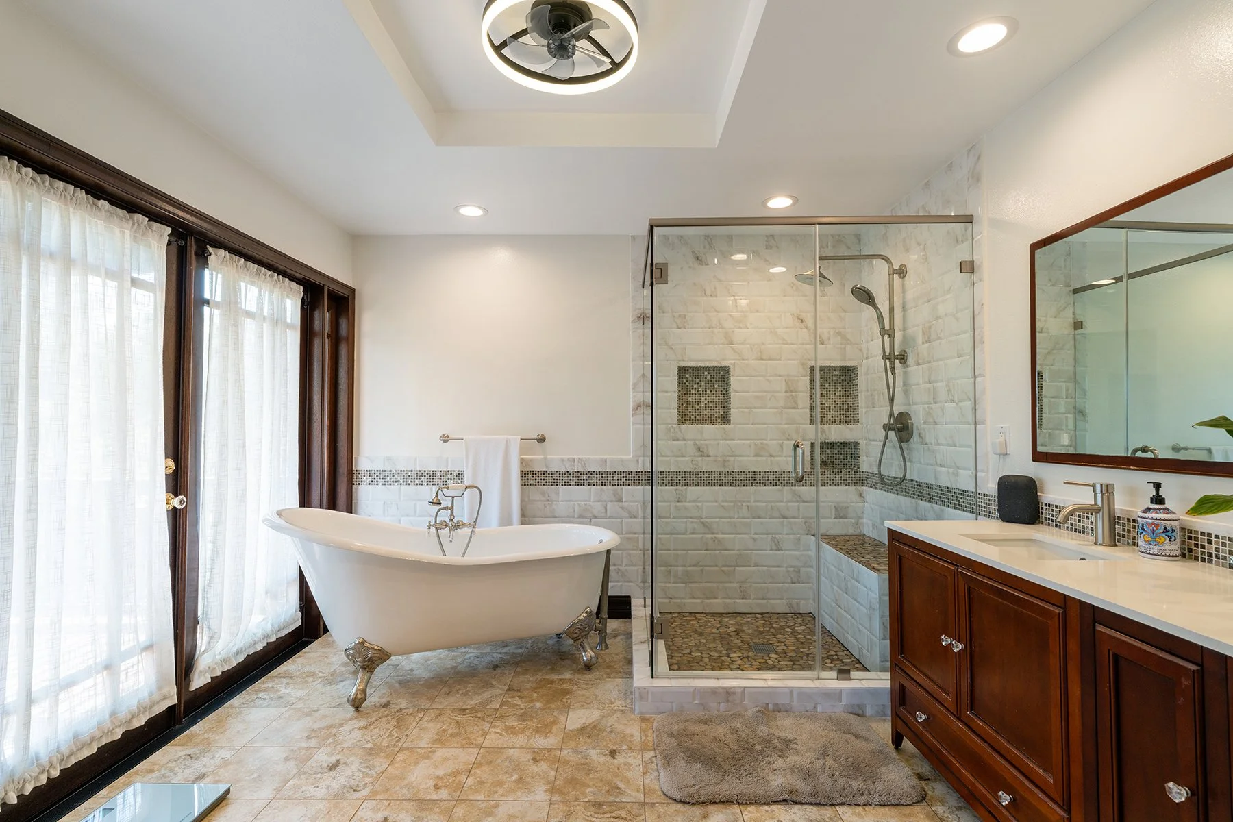 A bathroom with a clawfoot bathtub, a glass-enclosed shower, a wooden vanity with a sink, a large mirror, and a window with sheer curtains.