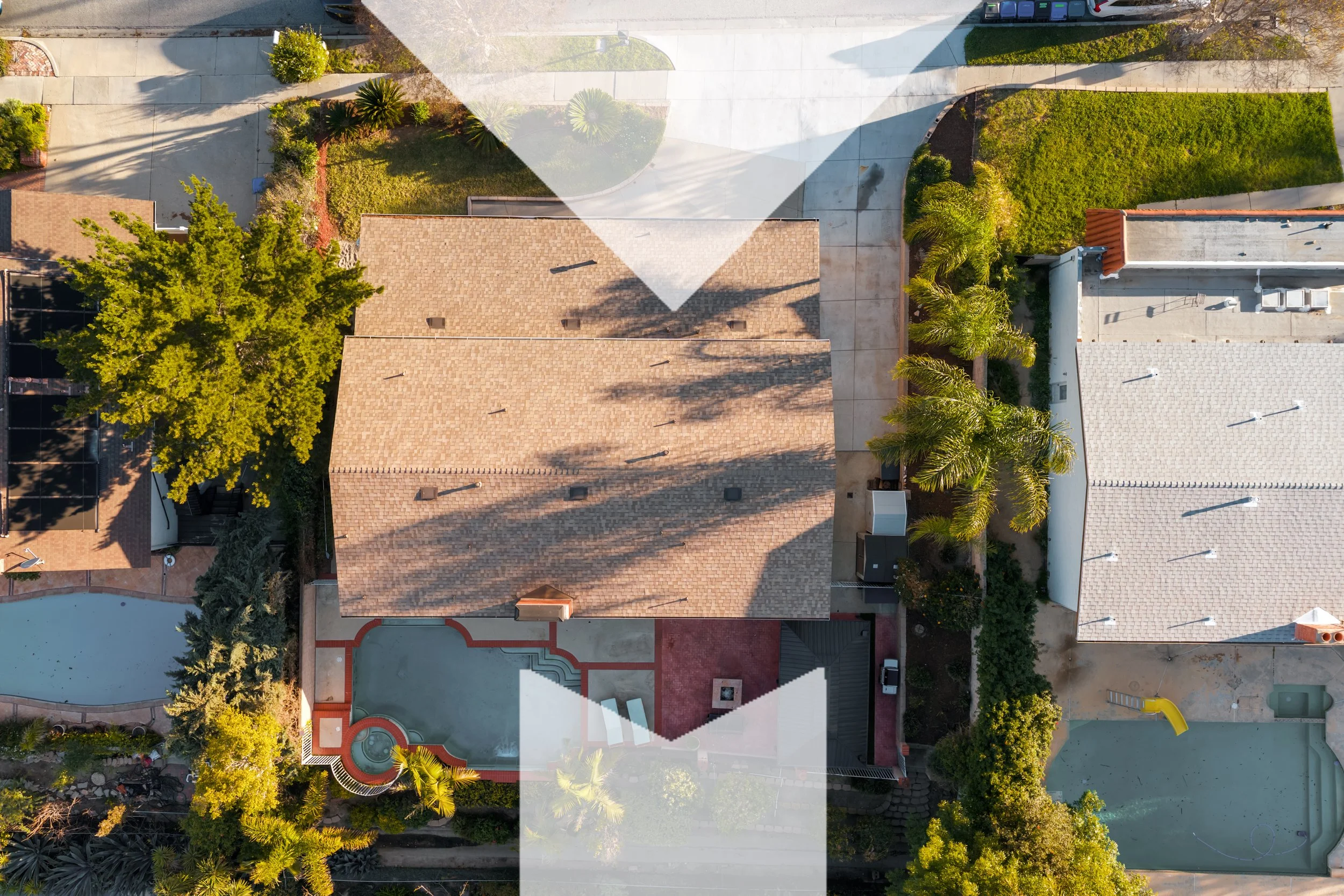 An aerial view of a residential backyard with two houses, a playground, a swimming pool, trees, shrubs, and lawns, with sunlight casting long shadows.