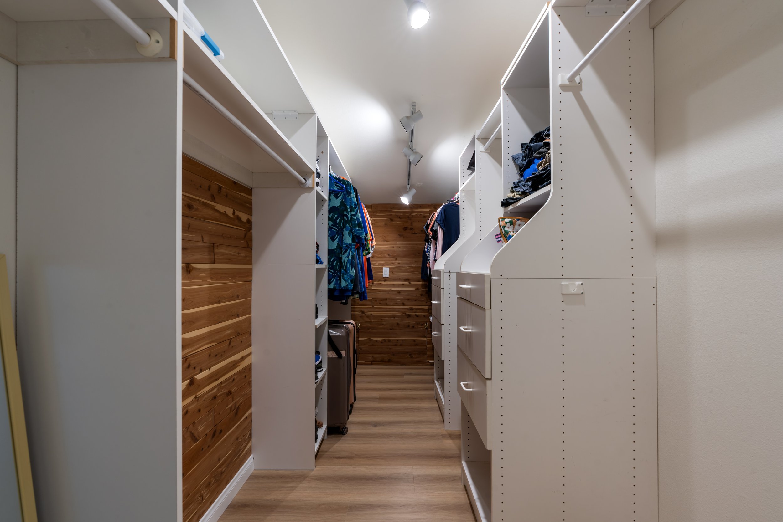A walk-in closet with white shelving units and hanging clothes, wooden accent wall, and wooden floor.