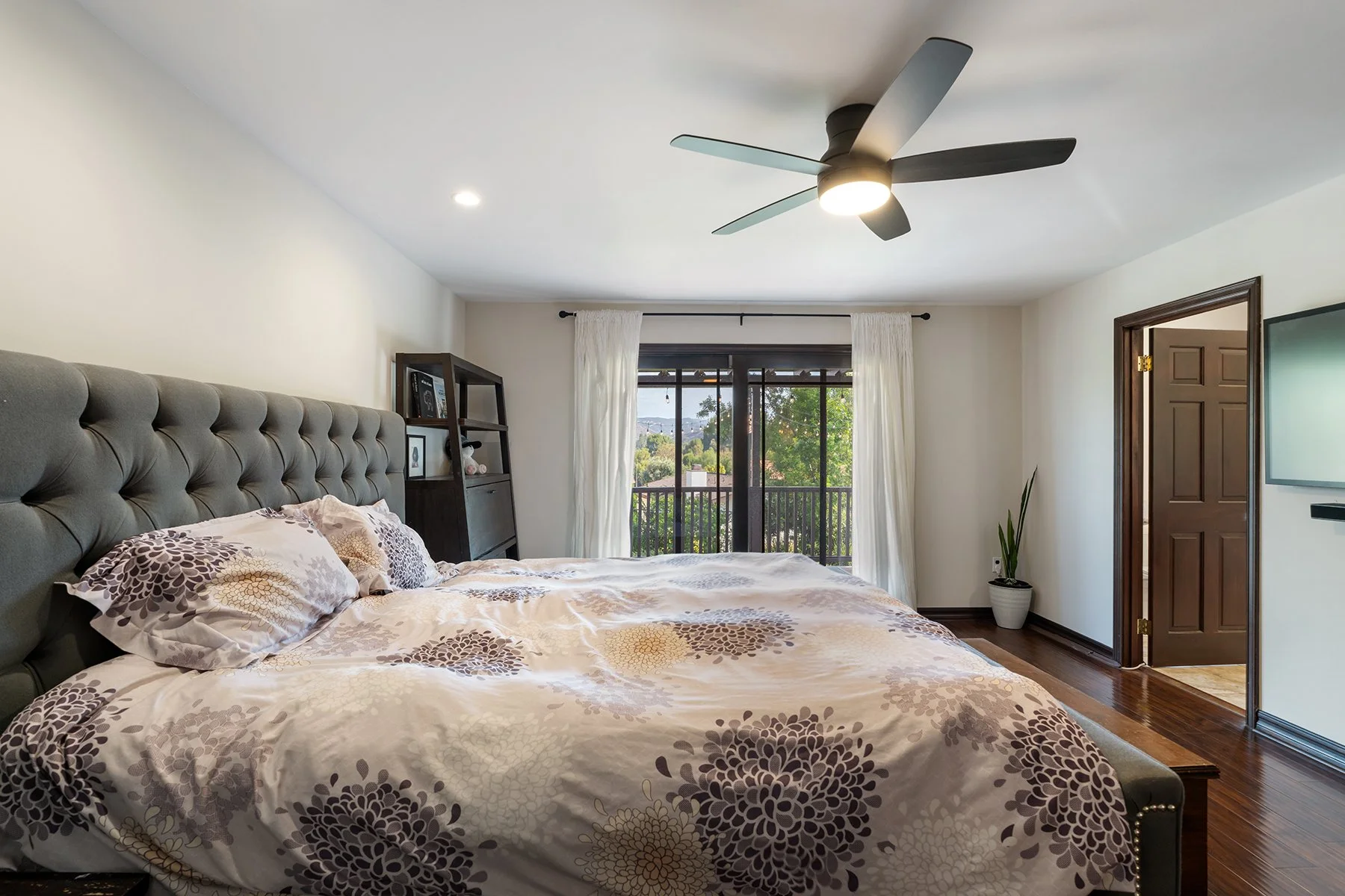 A bedroom with a large bed, tufted gray headboard, patterned bedding, and pillows. There is a window with white curtains, and a view of trees and buildings outside. A dark wooden shelf with framed photos and plush toys is next to the window. A ceiling fan with a light is above, and a doorway leads to another room. A potted plant is near the door.