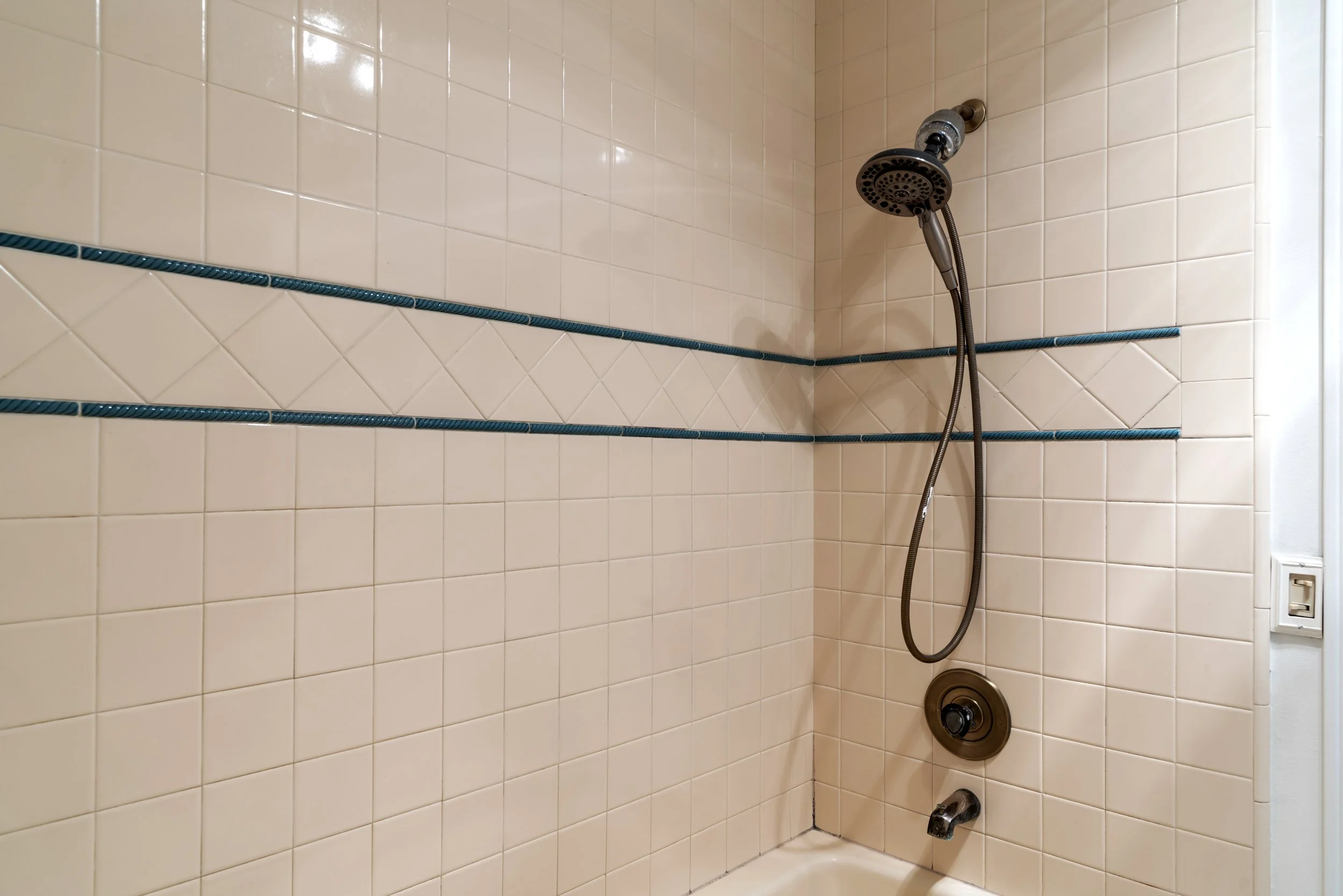 Shower with beige tiles, two horizontal blue decorative tiles, a showerhead, and a control knob.